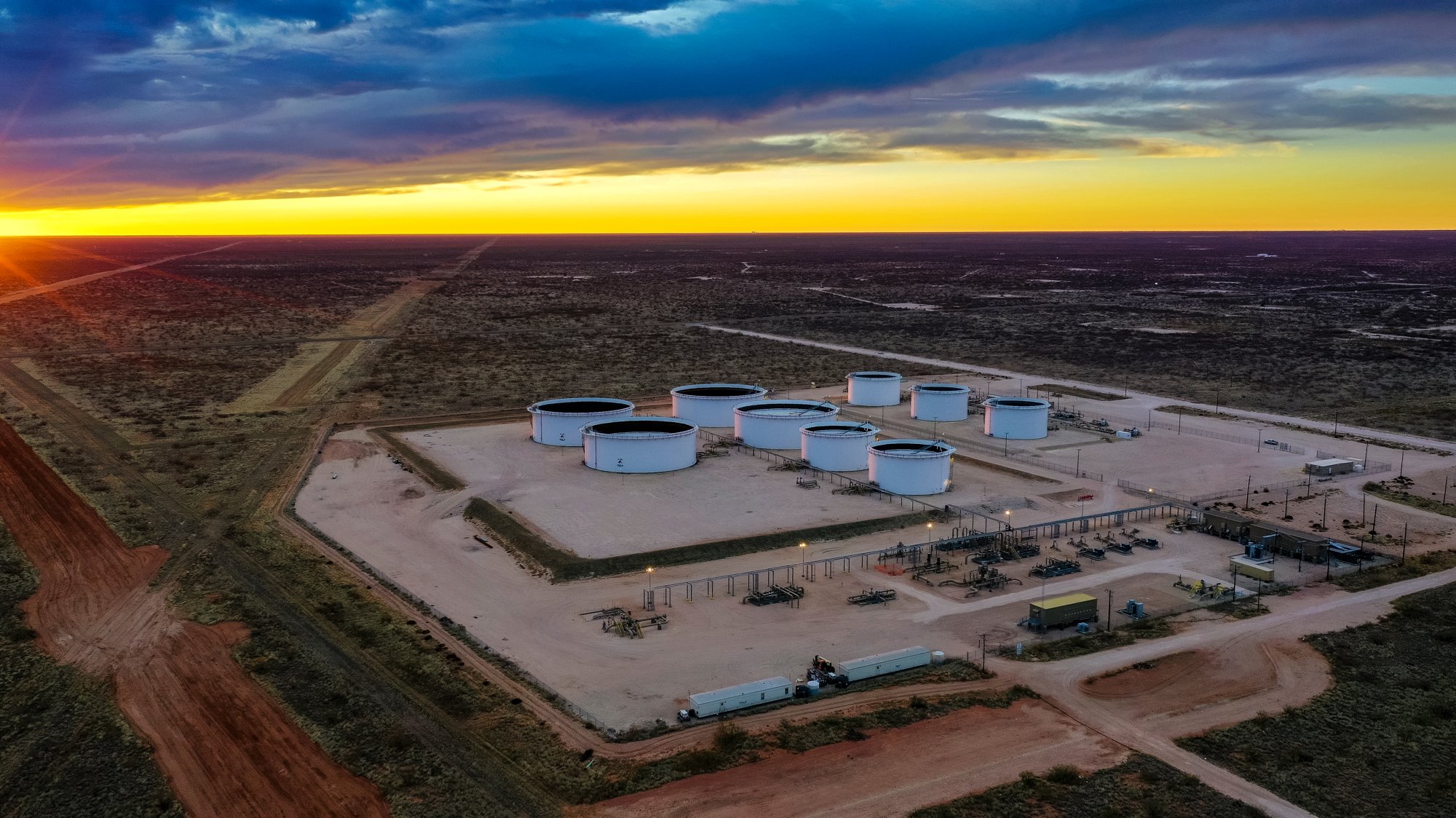 An oil or gas storage facility with multiple large white storage tanks in a desert landscape at sunset, with surrounding dirt roads and industrial equipment.
