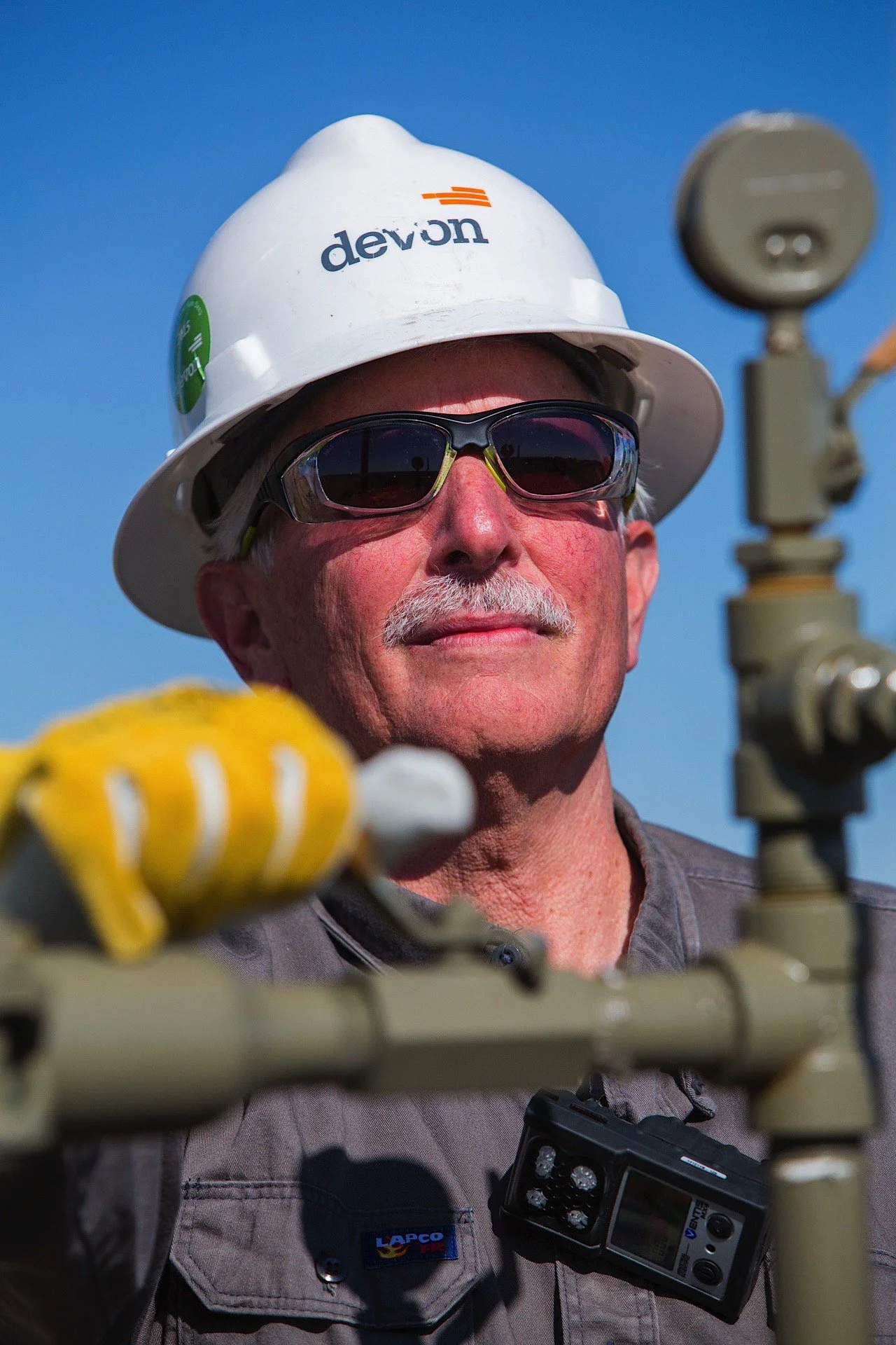 A man wearing a white hard hat, sunglasses, and work gloves operating equipment outdoors against a blue sky.