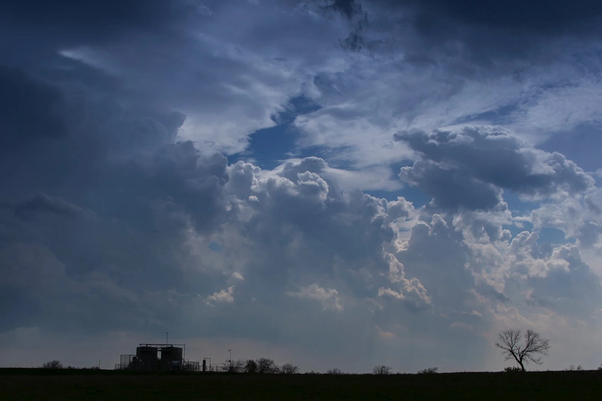 Empty field with a small building and a lone tree under a dramatic cloudy sky.