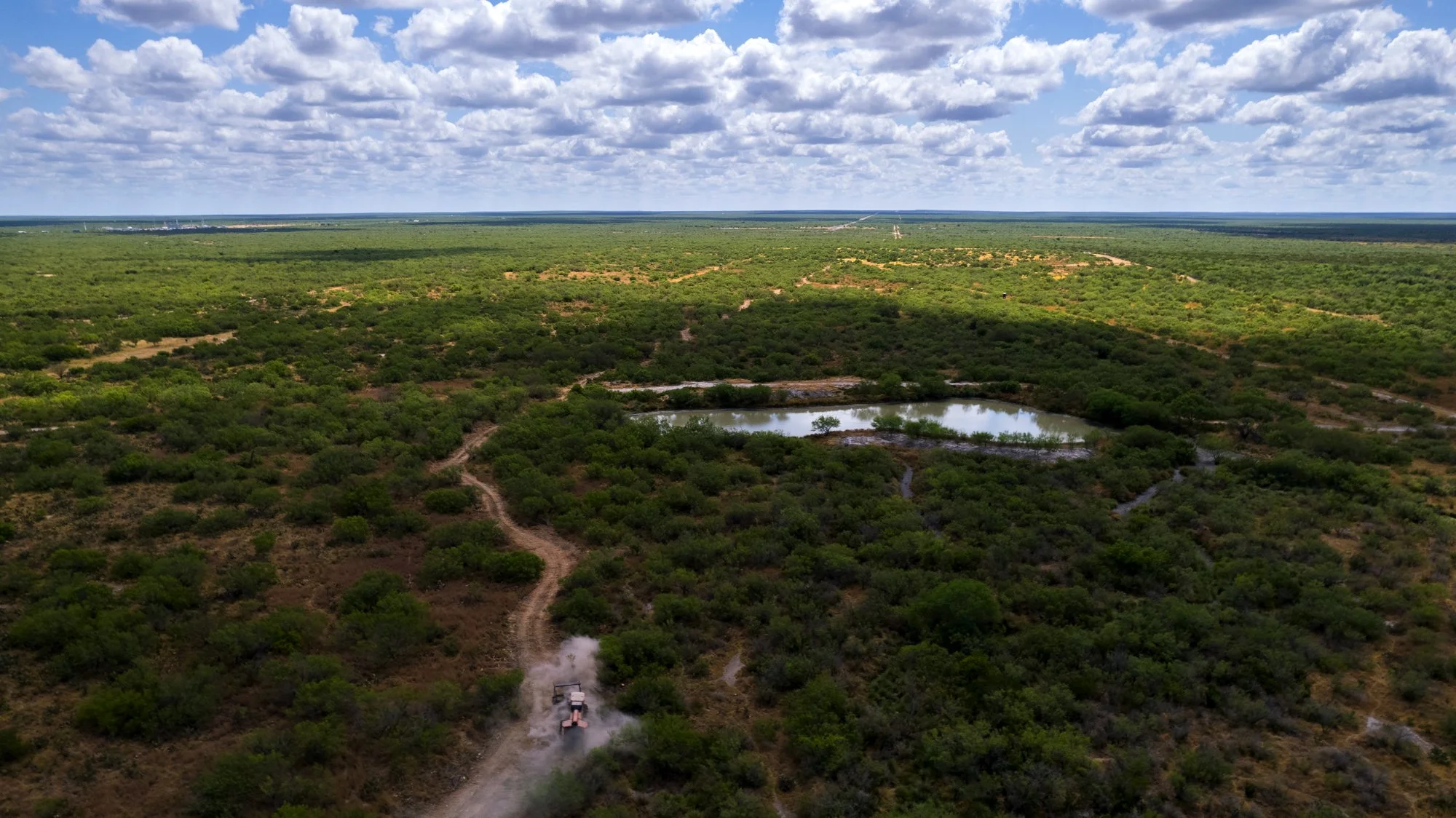 Aerial view of a vast green landscape with scattered shrubs and trees, a small lake, and a dirt trail with a vehicle kicking up dust.
