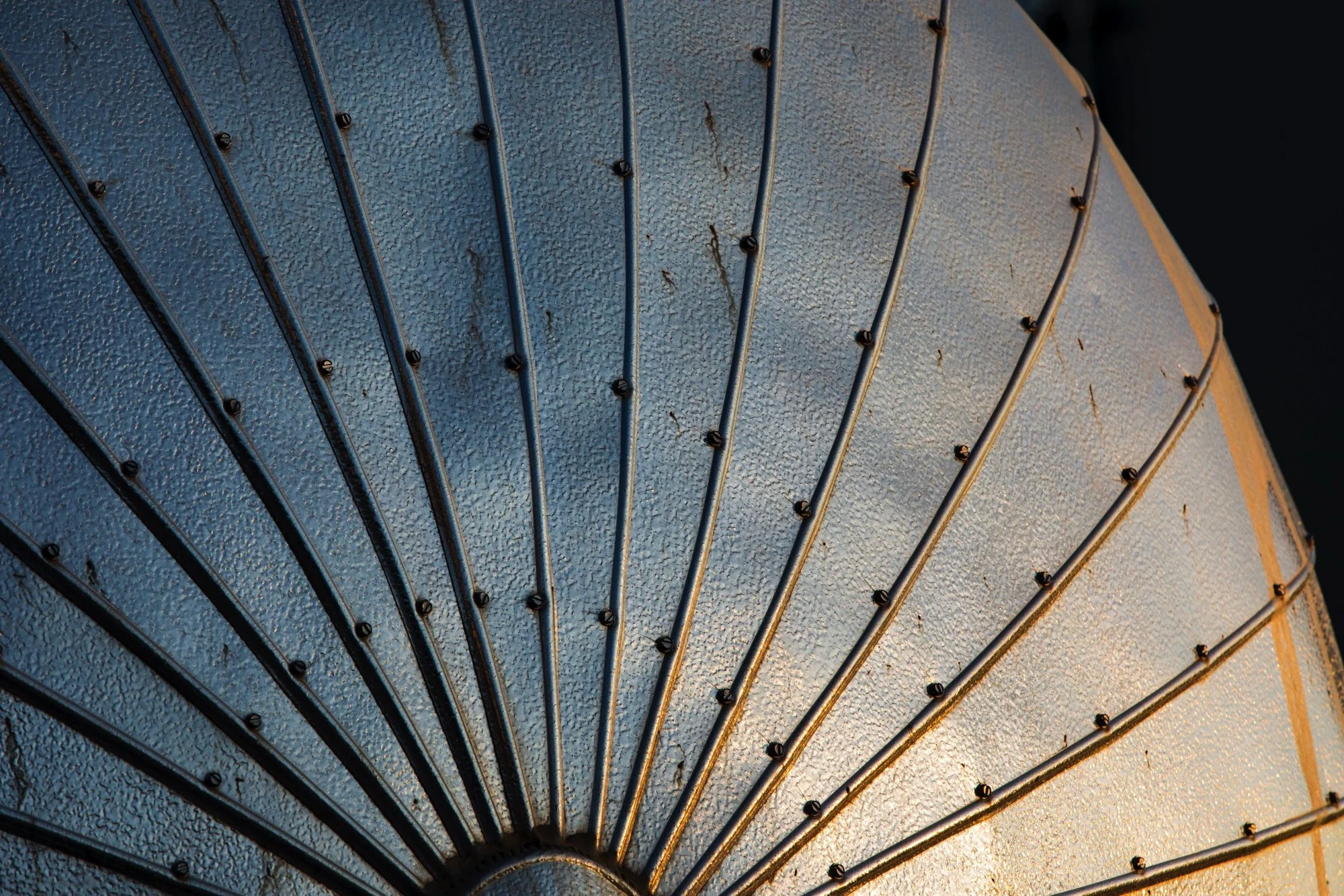 Close-up of a metallic, textured sunshade or umbrella with radiating metal ribs and small screws, illuminated by warm sunlight.