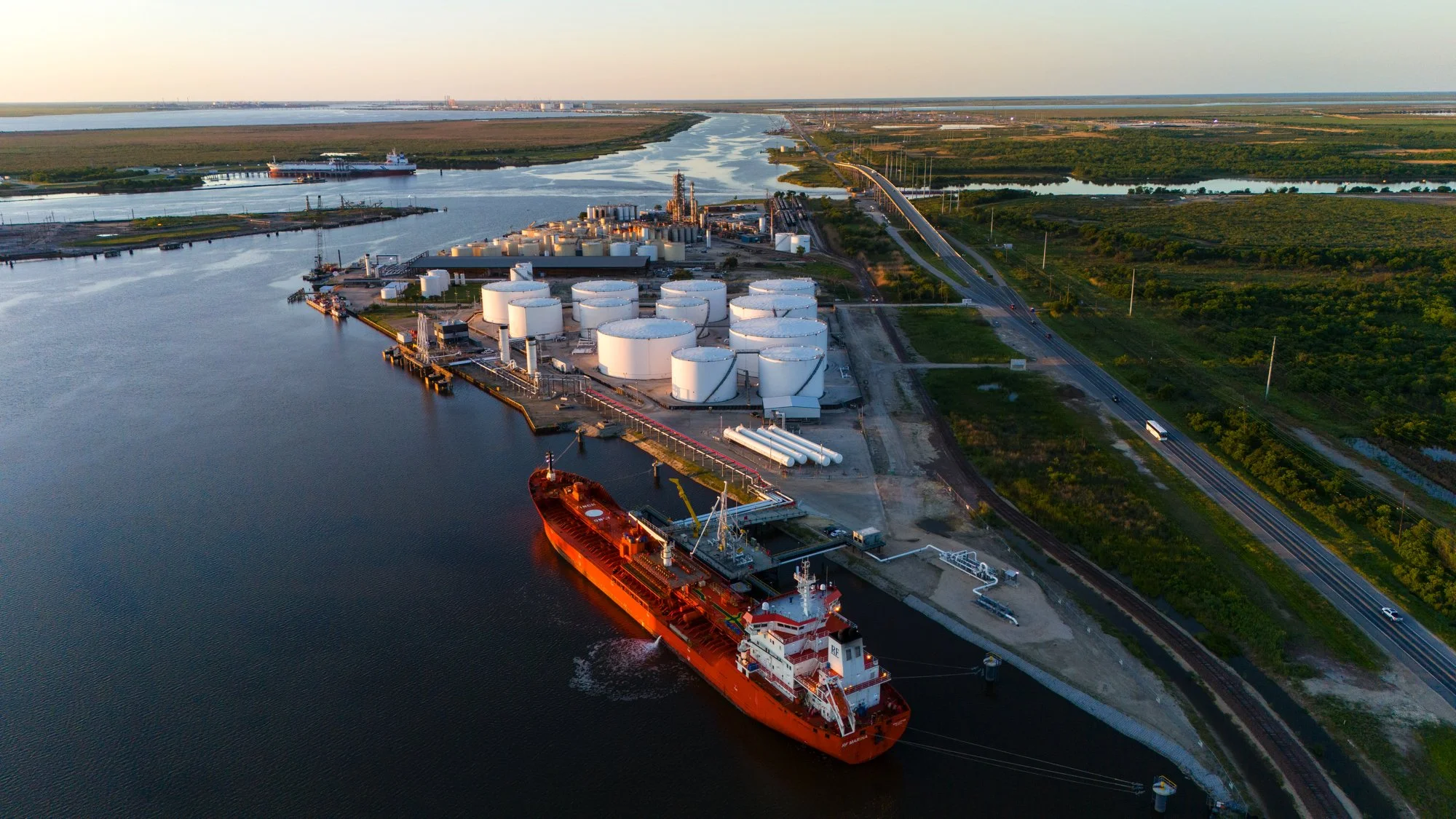 A large orange tanker ship docked at an industrial port with several white storage tanks and pipelines, surrounded by water, roads, and lush green land.