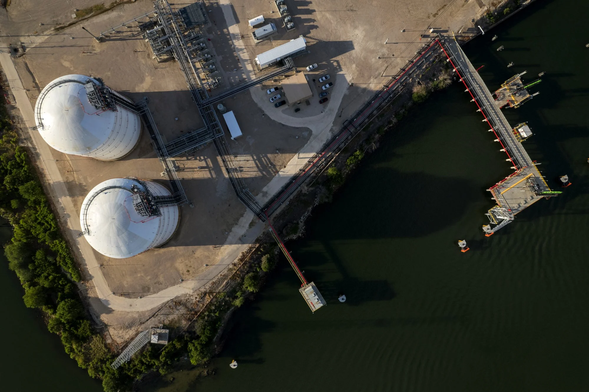 An aerial view of an industrial facility by a body of water, with two large white storage tanks, interconnected pipes, and a foghorn on a small pier extending into the water.