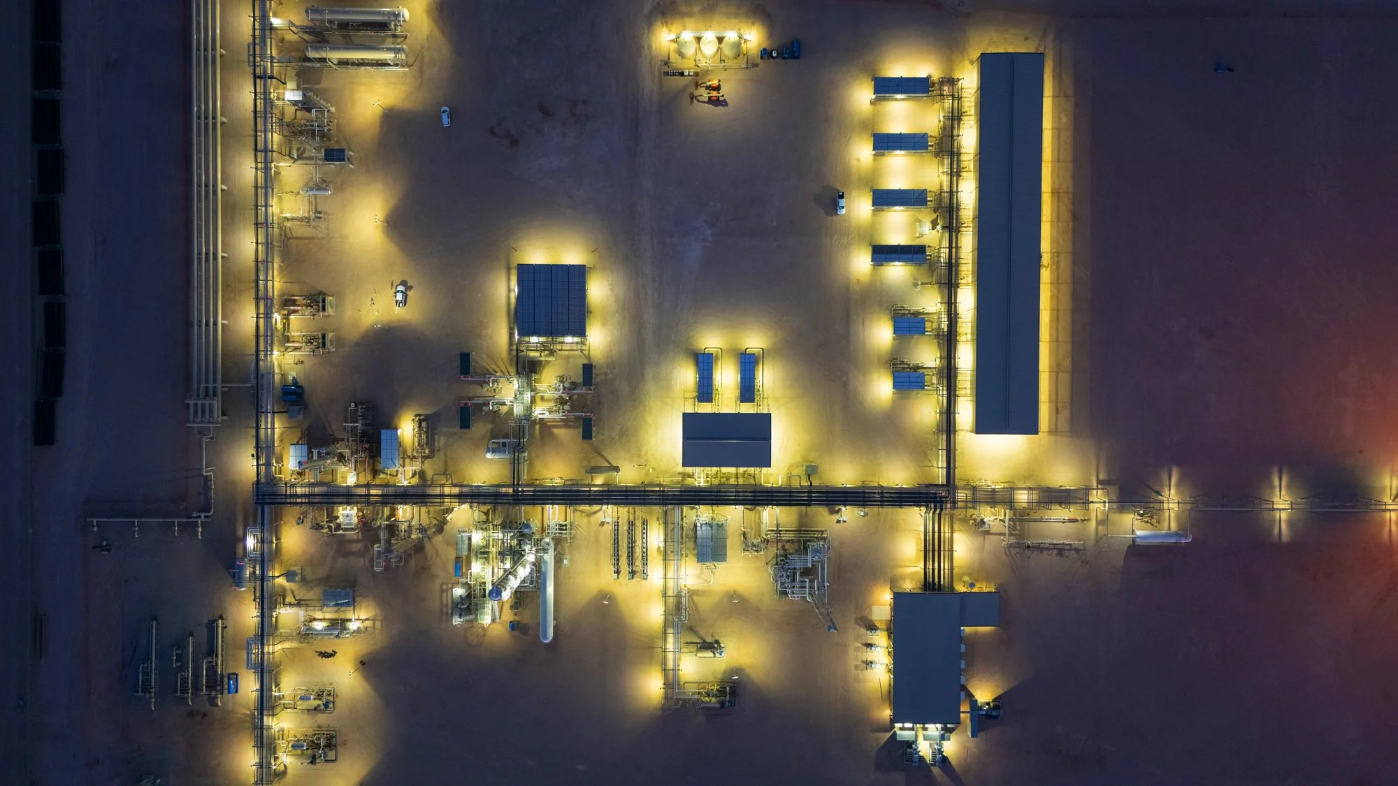 An aerial view of a solar power facility at night with multiple solar panels, structures, and pipelines illuminated by yellow lights.
