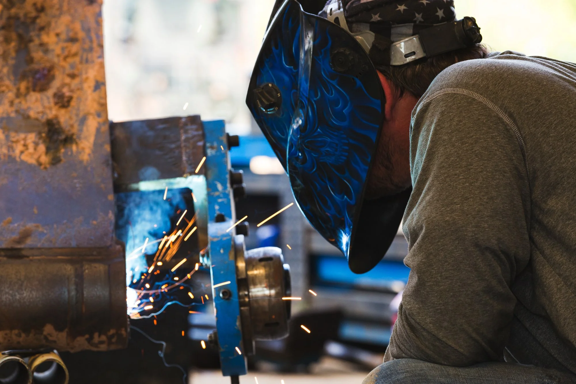A man welding metal while wearing a protective helmet with a blue flame design, sparks flying from the welding process.