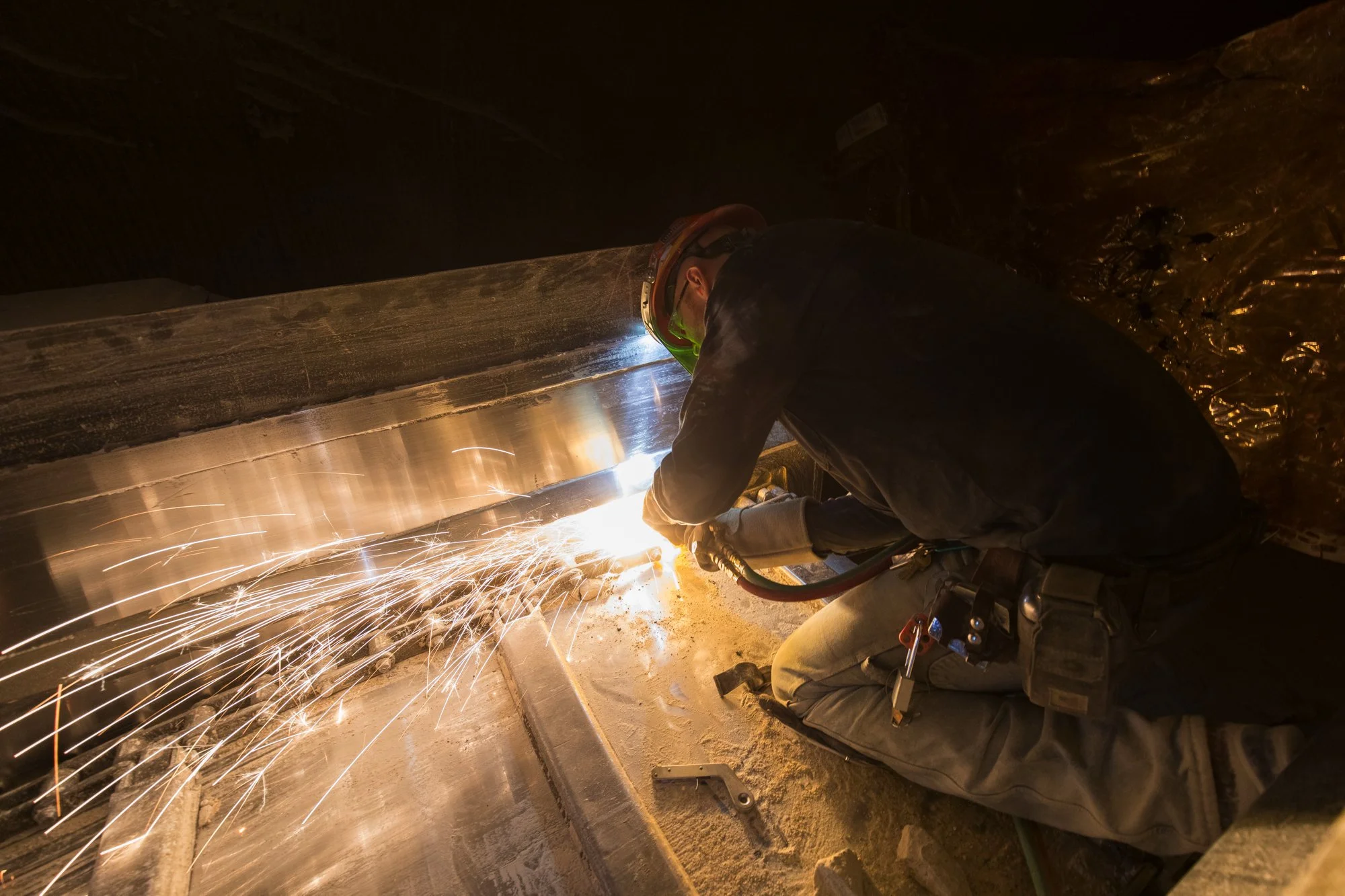 Worker welding metal beam, sparks flying, crouched on the floor, wearing protective gear and helmet, in an industrial setting.