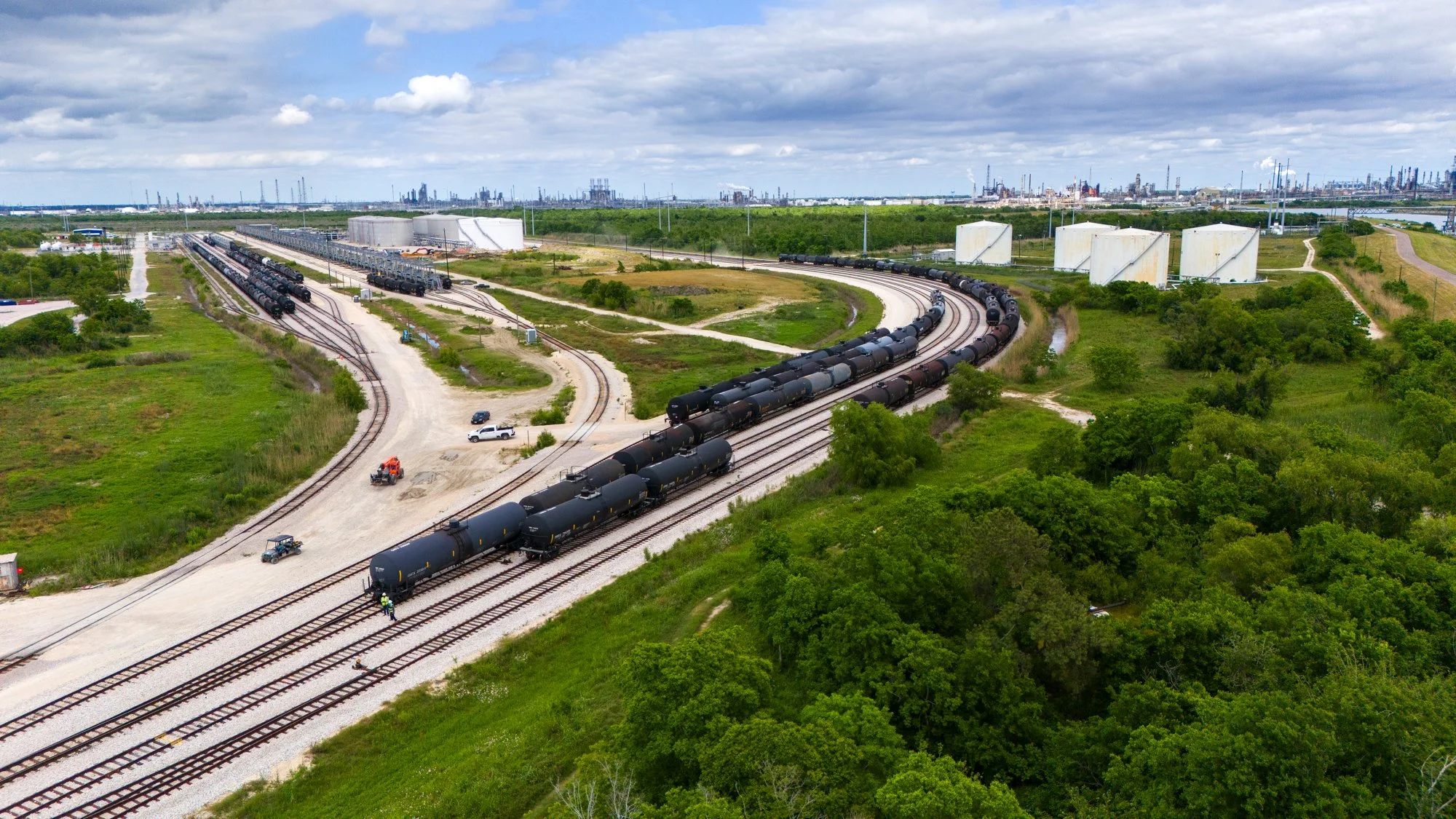 Aerial view of multiple black tanker train cars on curved railway tracks in a green industrial area with storage tanks and distant factory buildings.