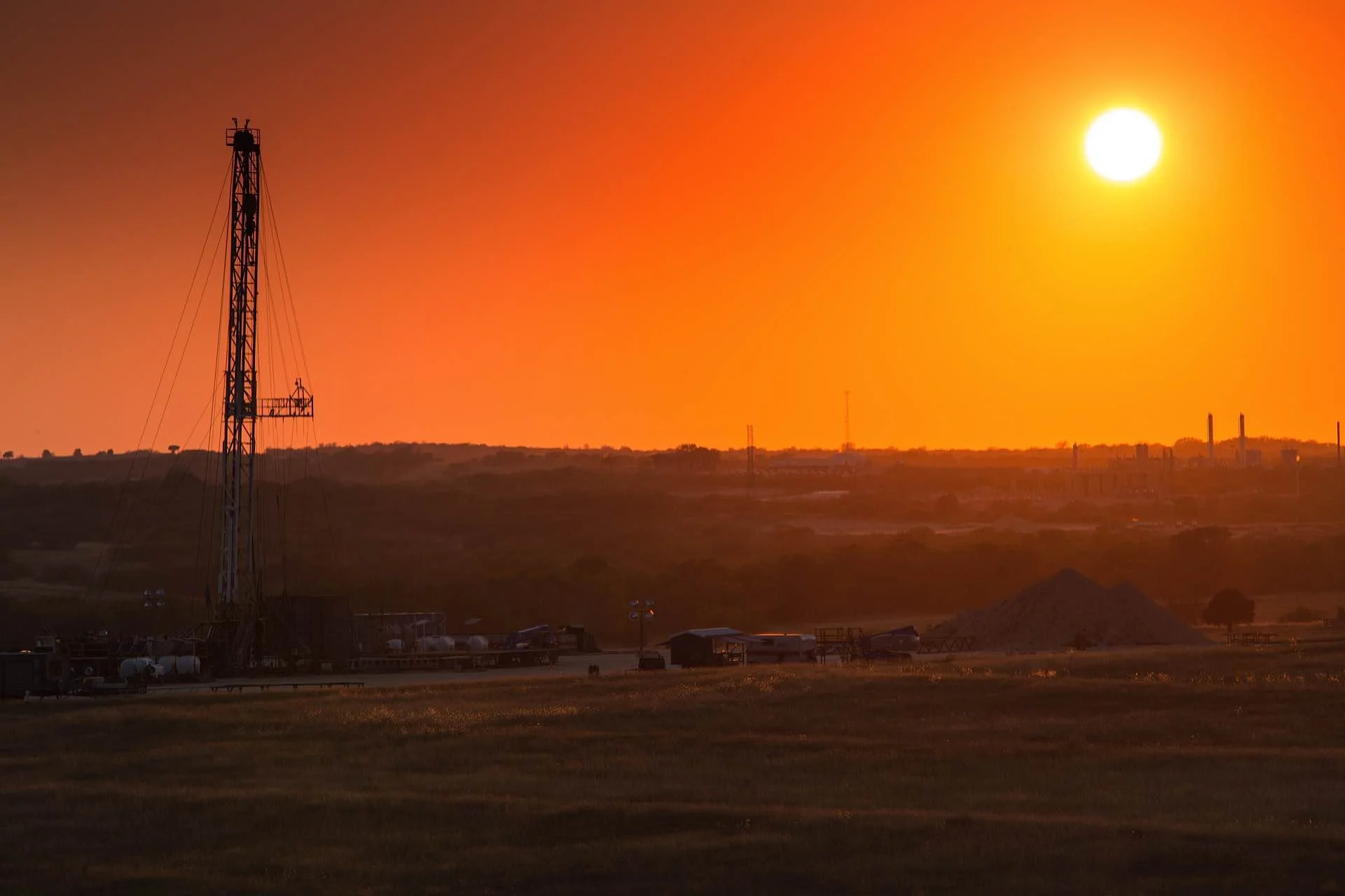 An oil rig in an open field during sunset with a bright sun in the sky and a few structures in the distance.