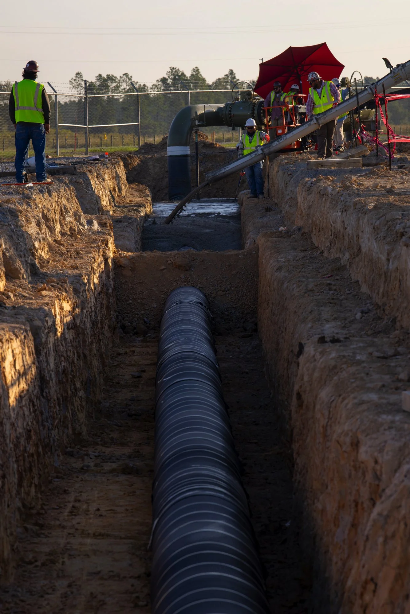 Construction workers install large pipeline pipes in a trench, with equipment and safety gear, under bright sunlight.