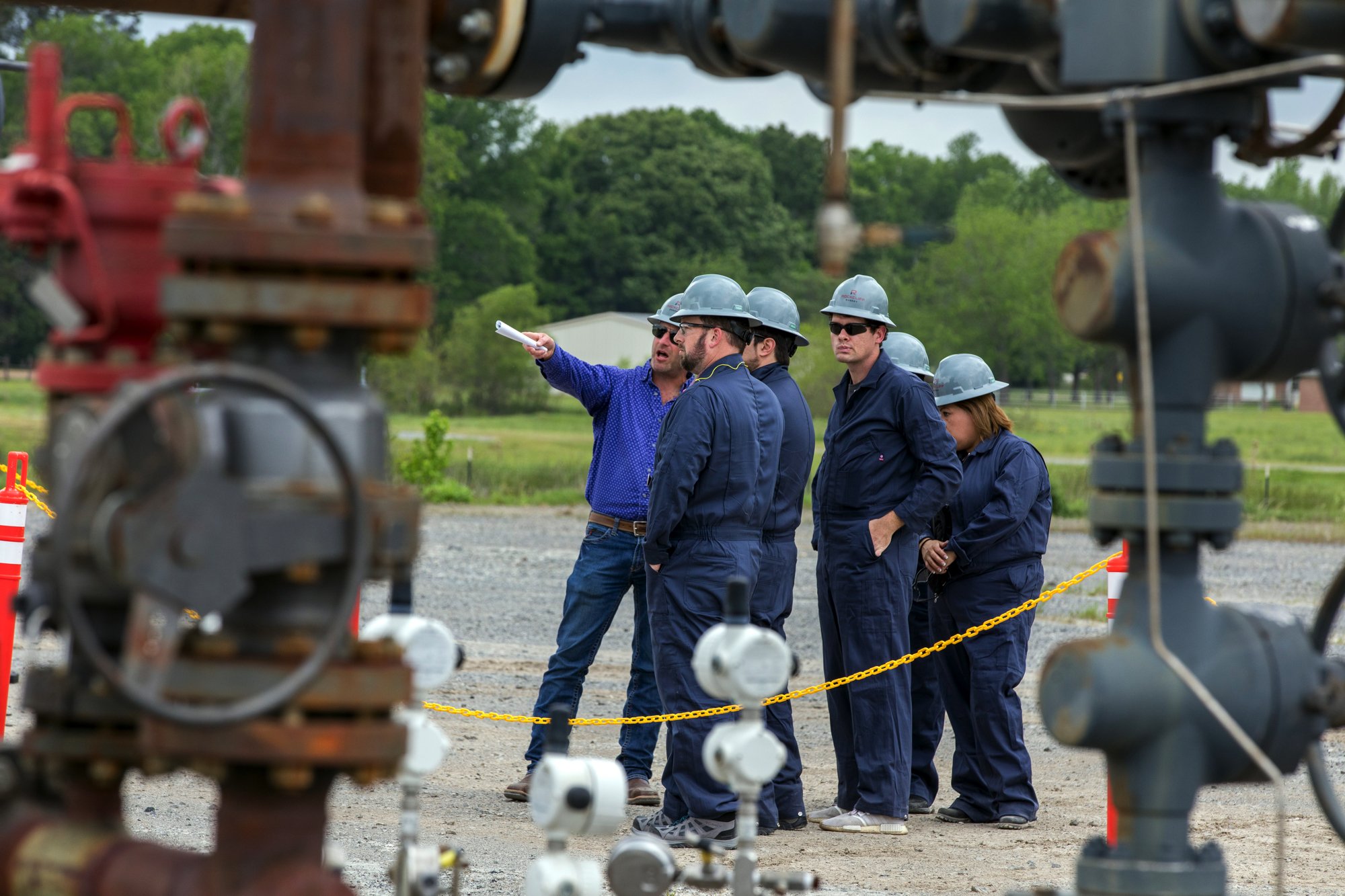 Group of workers wearing safety helmets and dark blue uniforms inspecting industrial equipment outdoors, with a man in a blue shirt pointing and holding a rolled document, surrounded by safety barriers.