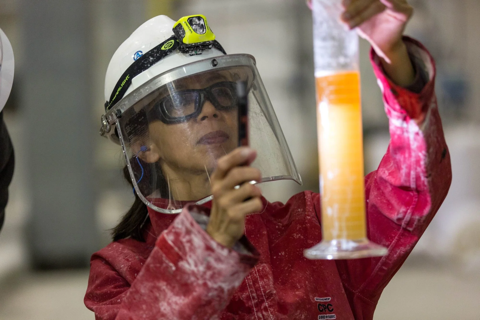 A woman wearing a white safety helmet, goggles, and a red protective jacket holding a measuring instrument in an industrial setting.