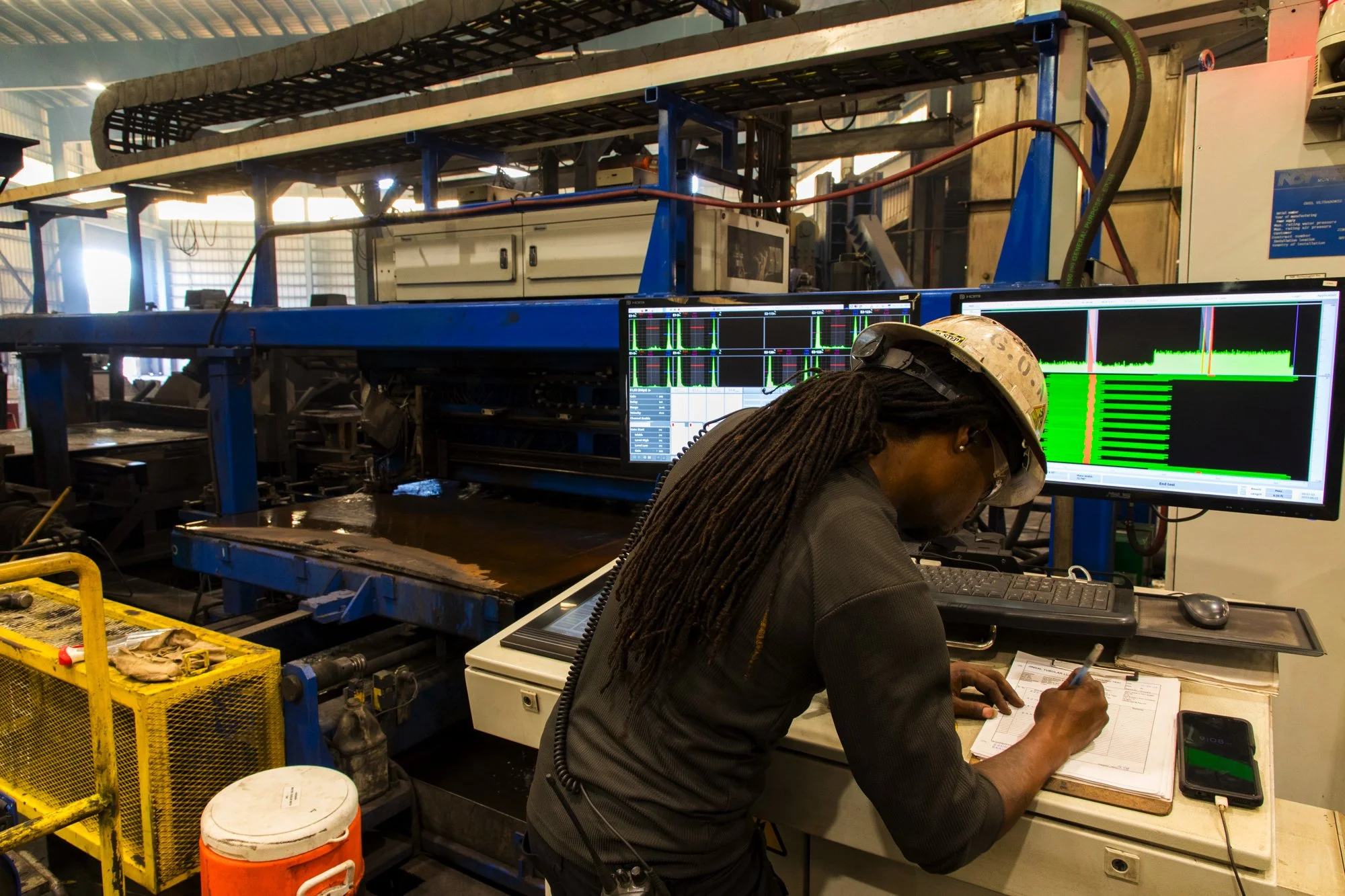 A worker in a hard hat operating industrial equipment with two computer monitors displaying data and graphs in a factory setting.