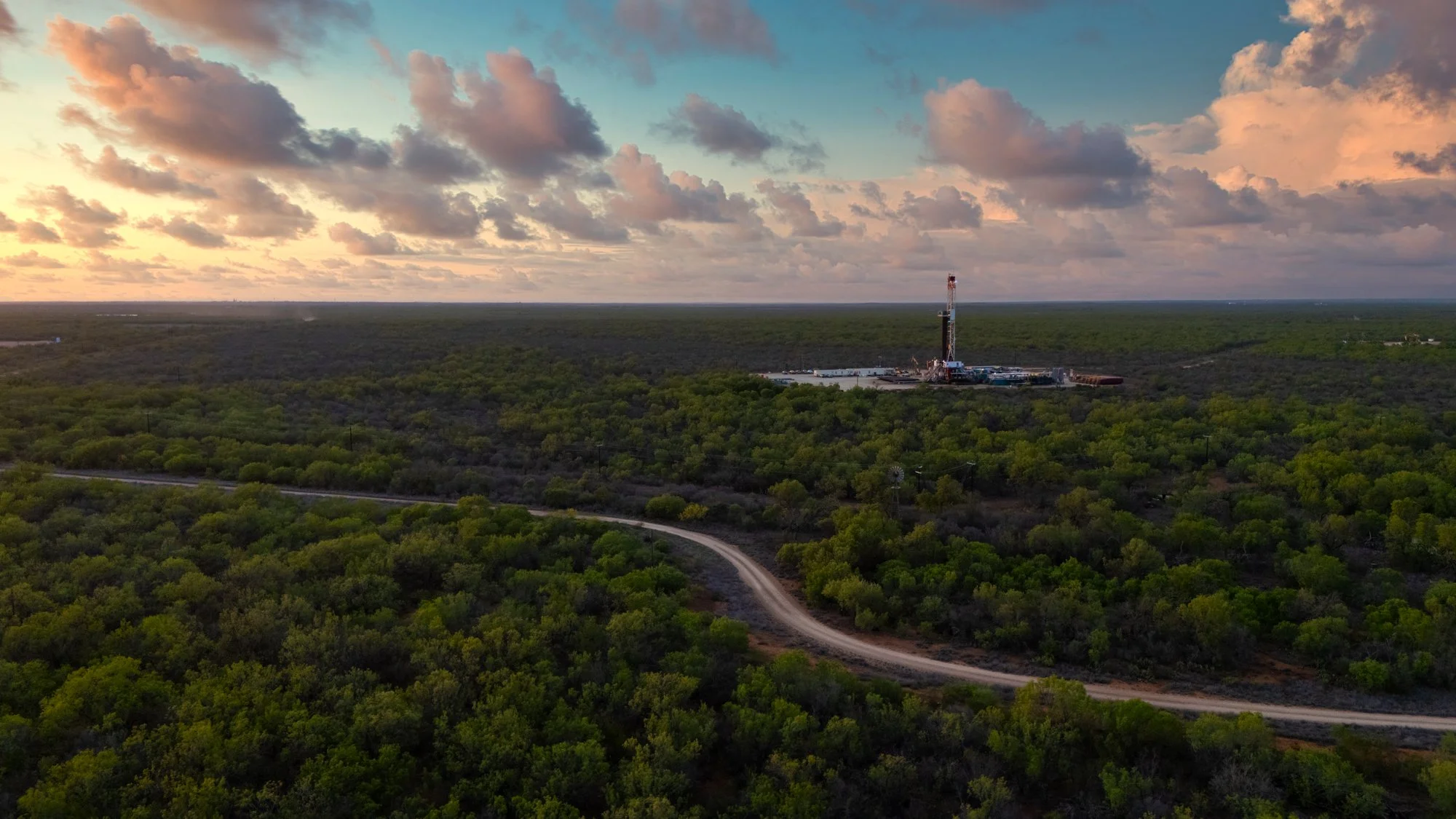 An aerial view of a drilling rig in a green forested area at sunset, with a dirt road winding through the trees and a partly cloudy sky overhead.