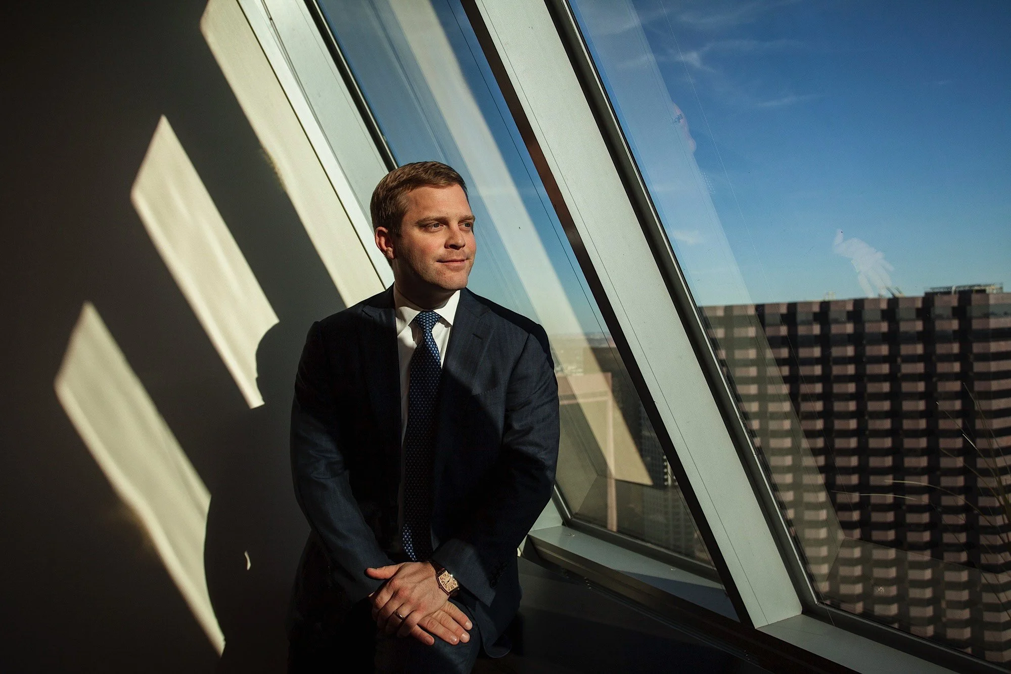 A man in a dark suit and tie standing by a large, angled window in a high-rise building, gazing out at the city skyline on a sunny day.