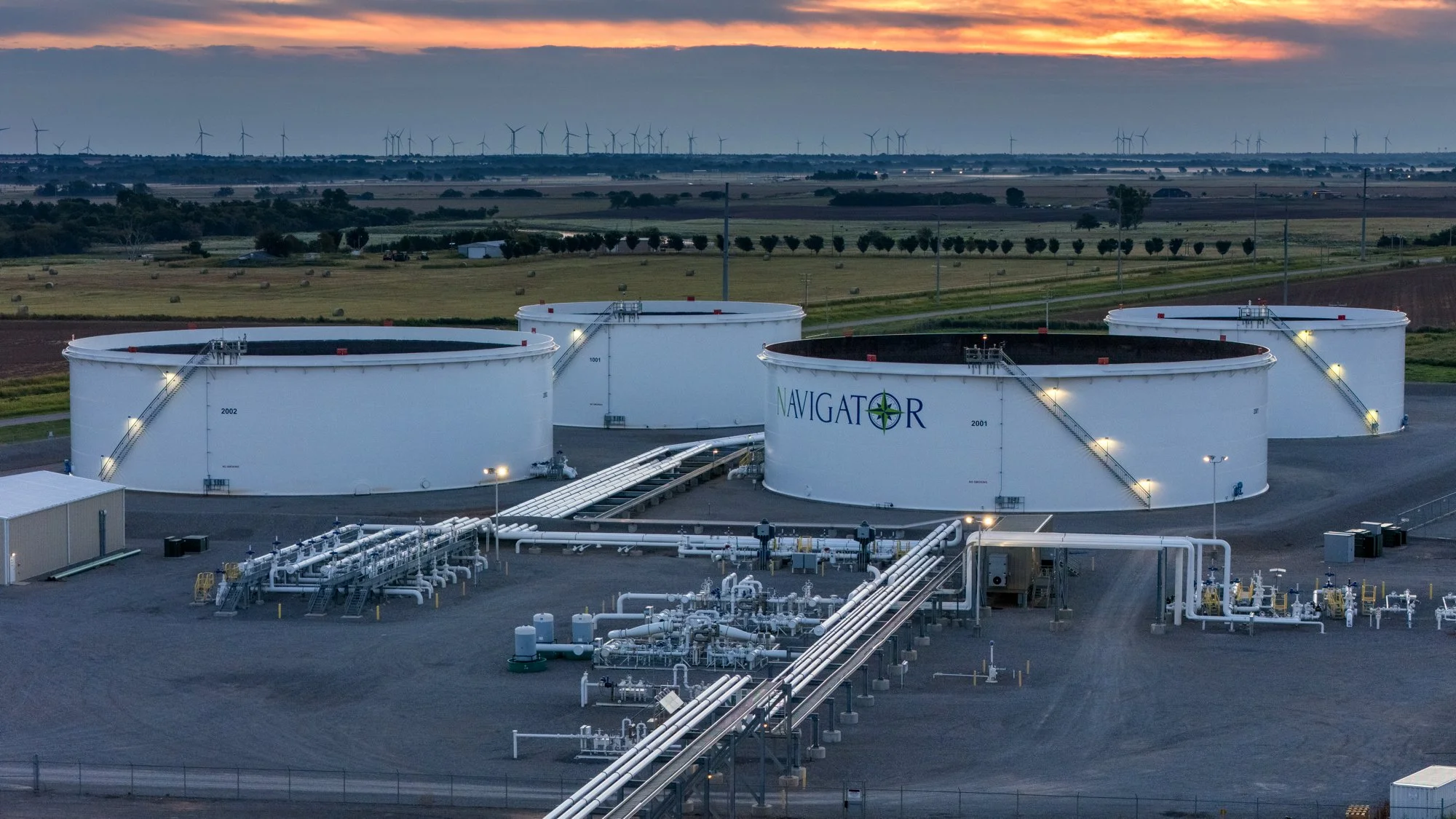 Large white fuel storage tanks labeled Navigator with pipelines and industrial equipment in foreground, rural landscape with wind turbines in background at dusk.
