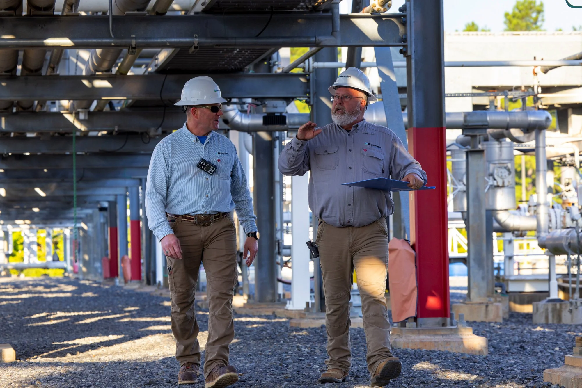 Two male industrial workers wearing safety helmets and work clothes, walking and talking at an industrial site with pipes and equipment in the background.