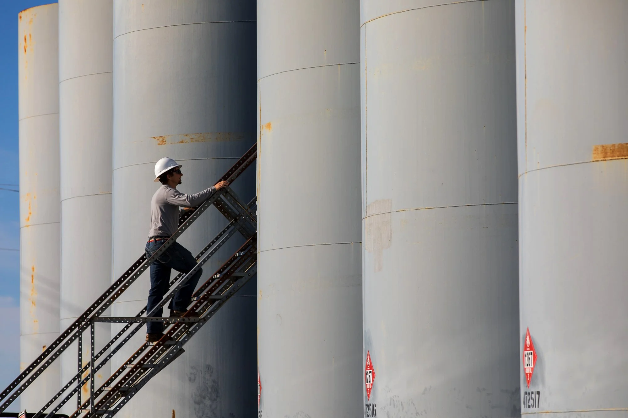 A person wearing a white safety helmet, sunglasses, and a gray shirt climbing a metal staircase next to large gray industrial storage tanks.