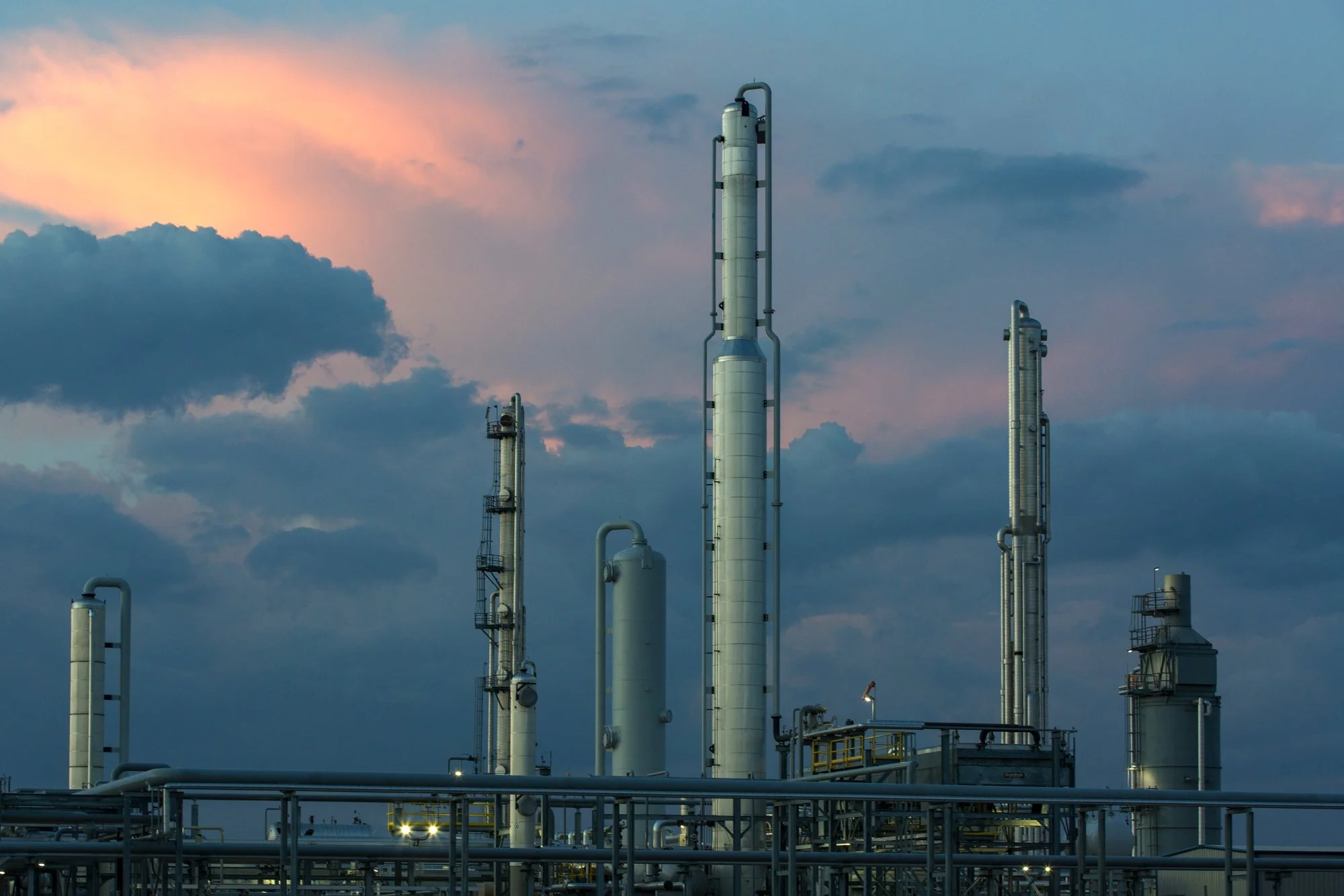 An industrial refinery with multiple tall metallic towers and pipes against a cloudy sky at dusk.