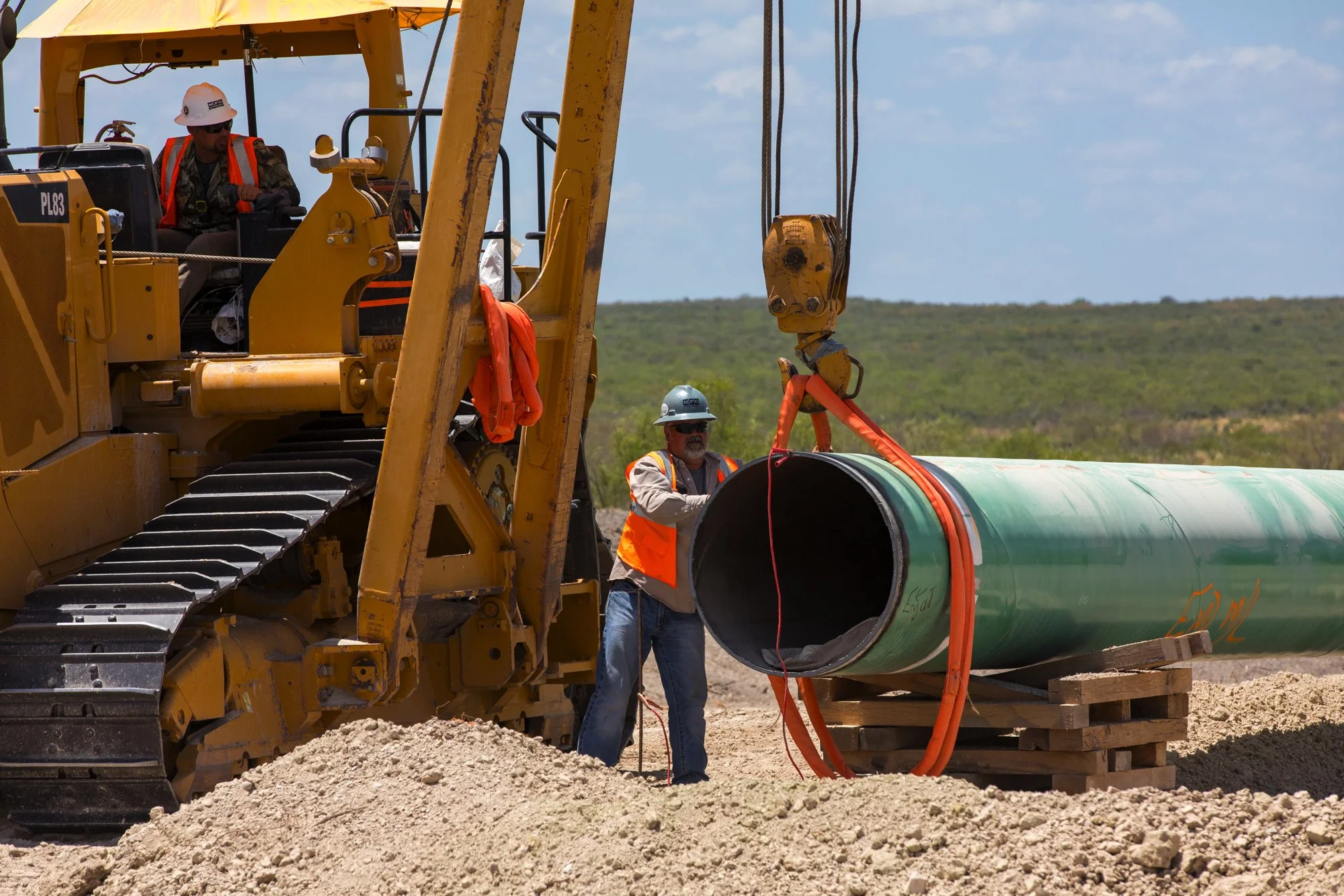 Construction workers installing a large green pipe using heavy equipment on a dirt site with a landscape background.