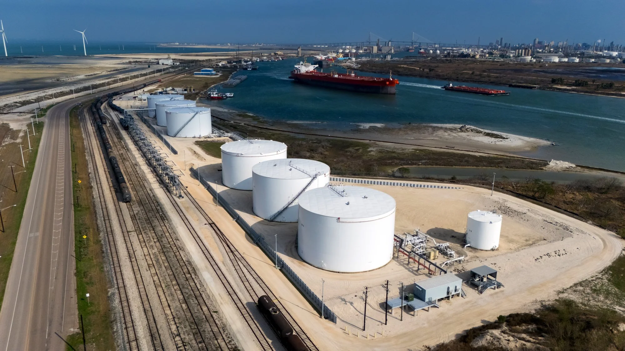 Aerial view of oil storage tanks near a waterway with ships and a city skyline in the background.