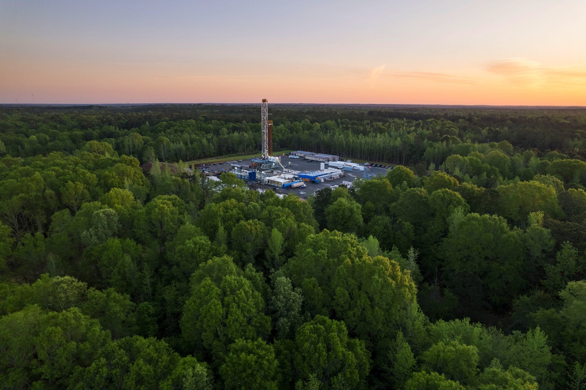 Aerial view of a drilling rig in a forested area during sunset.