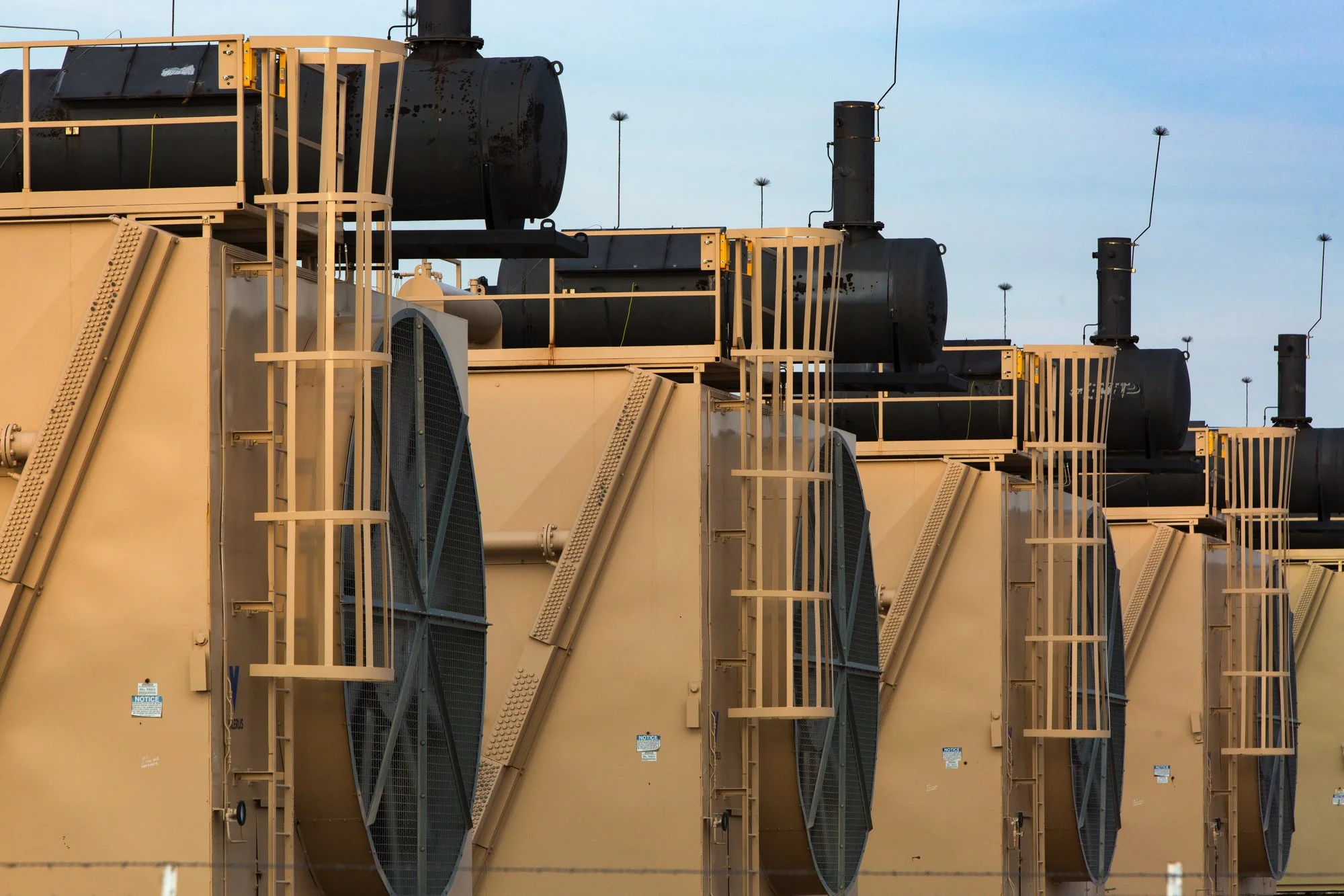Row of large industrial air conditioning units with fans and exhaust pipes outdoors, under a partly cloudy sky.