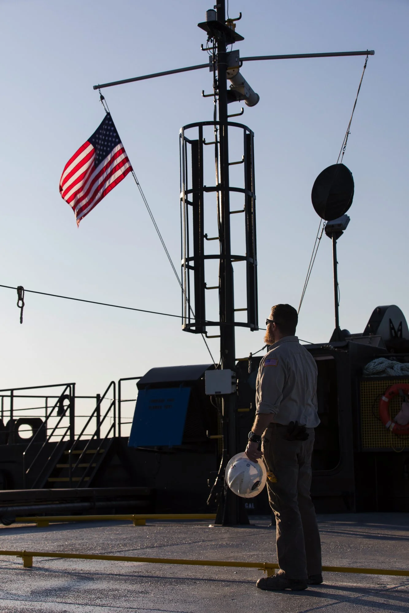 A crew member in uniform holding a white helmet with a ship's deck and mast with an American flag in the background.