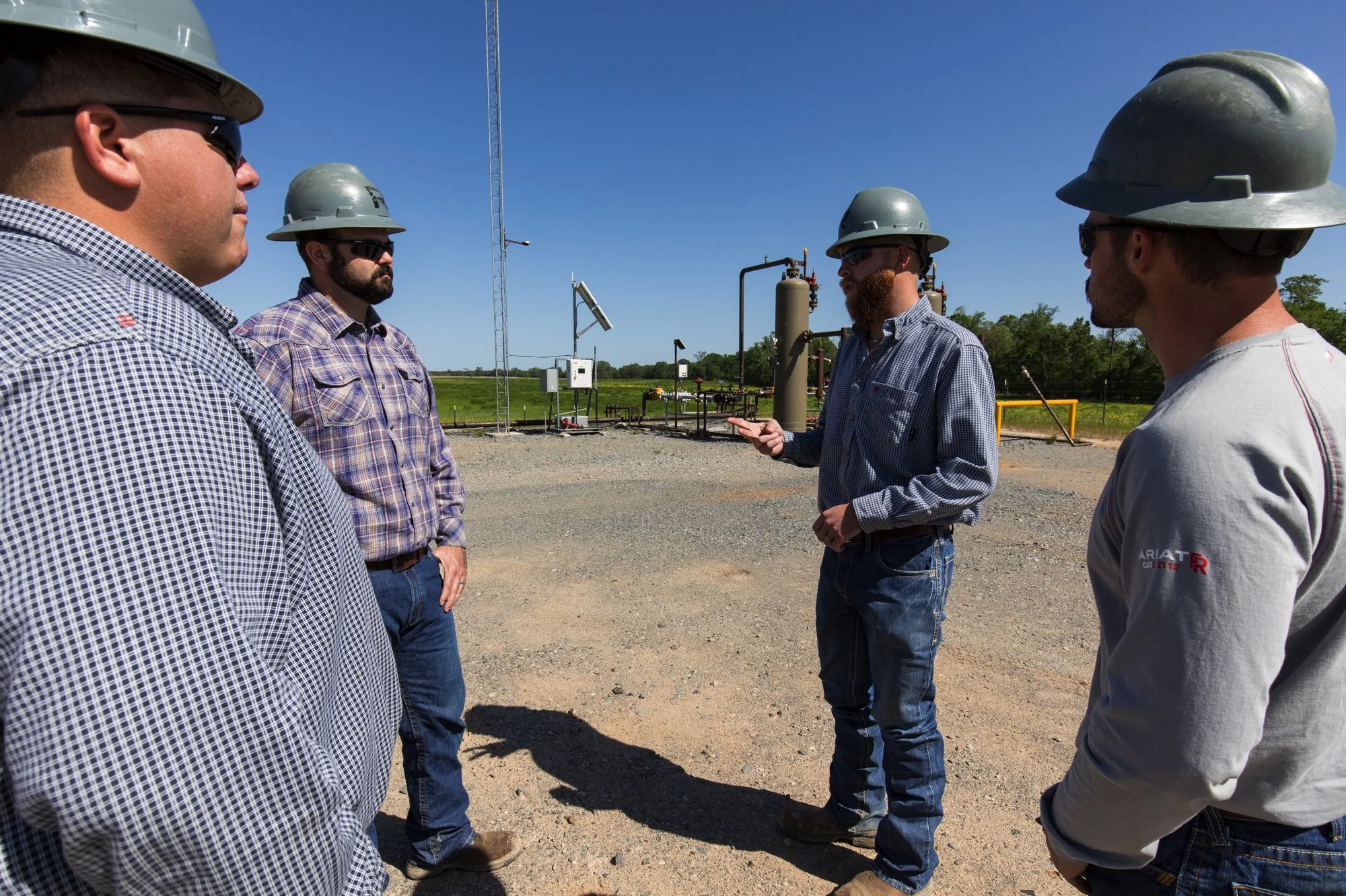 Four men wearing hard hats and casual work clothes standing outdoors in a gravel area with industrial equipment and a communications tower in the background, engaged in a discussion.