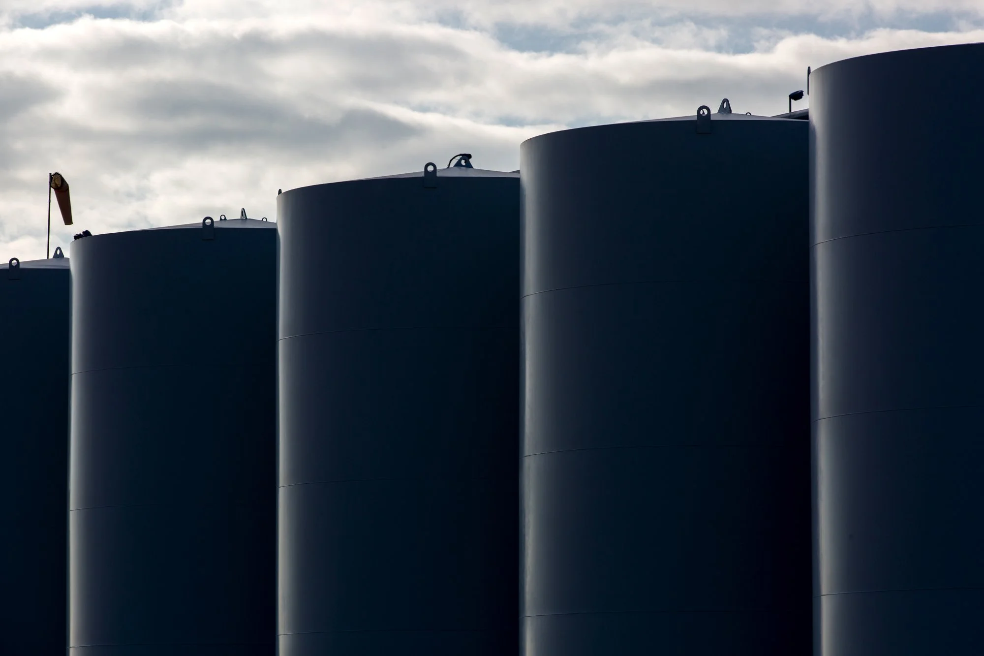 Close-up of large dark blue industrial storage tanks against cloudy sky.