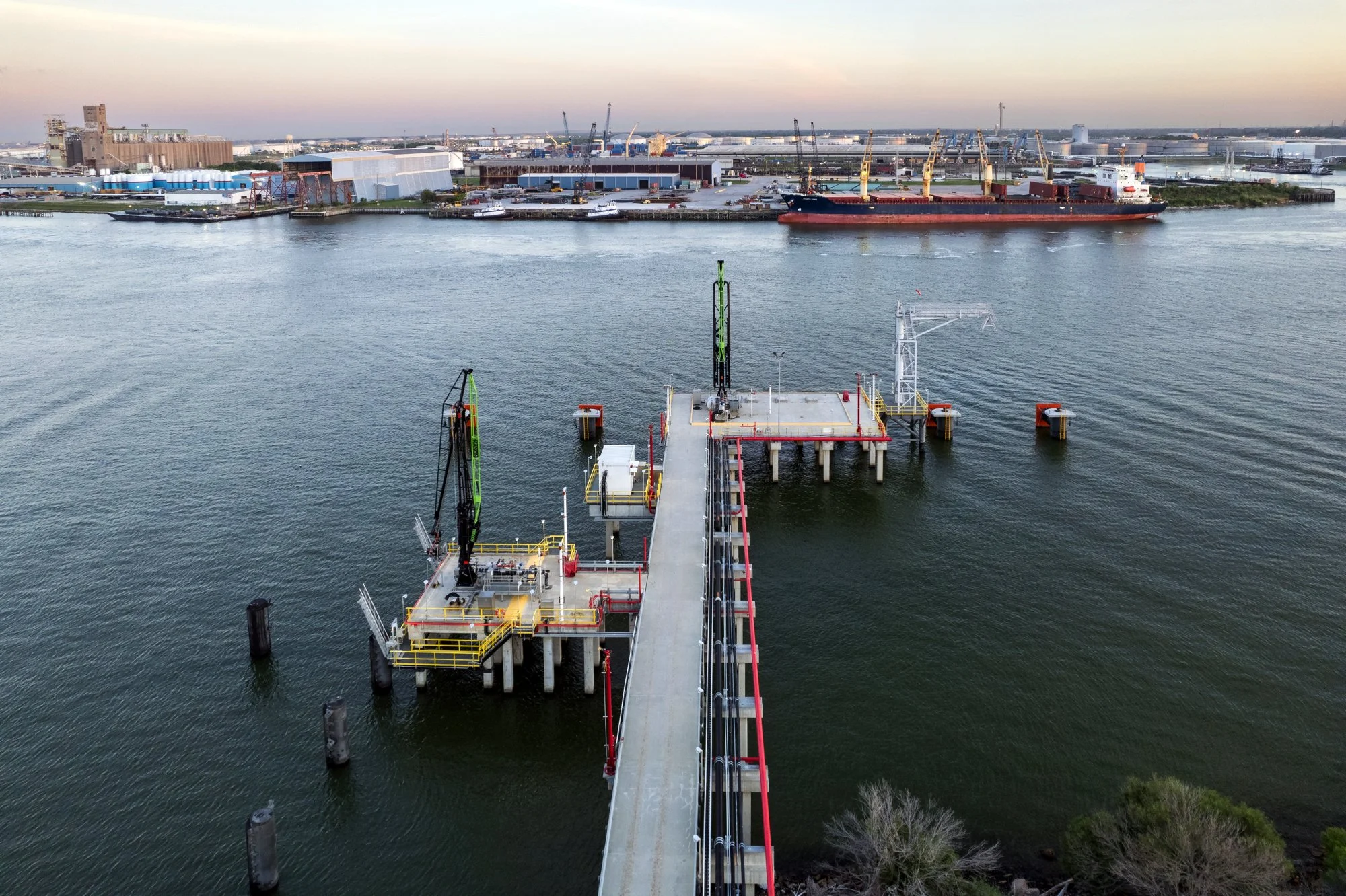 An aerial view of an offshore oil or gas drilling platform in a body of water with industrial port facilities, ships, and cranes in the background during sunset.