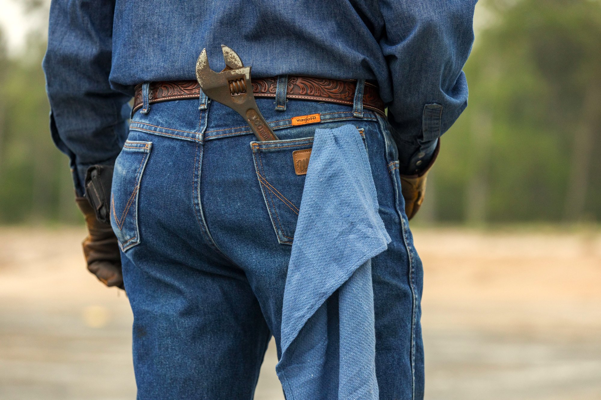 Back of a man wearing denim jeans, a dark shirt, a brown belt, and gloves, with a large adjustable wrench partially in his back pocket and a shirt hanging from his pocket.