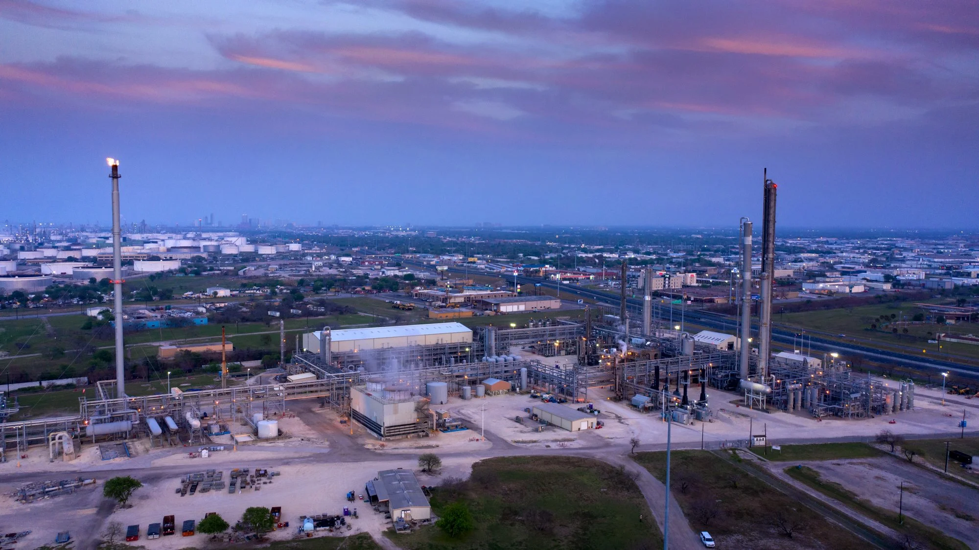 An industrial oil refinery with multiple tall distillation towers and storage tanks, situated in a flat landscape with highways and buildings in the background during dusk.
