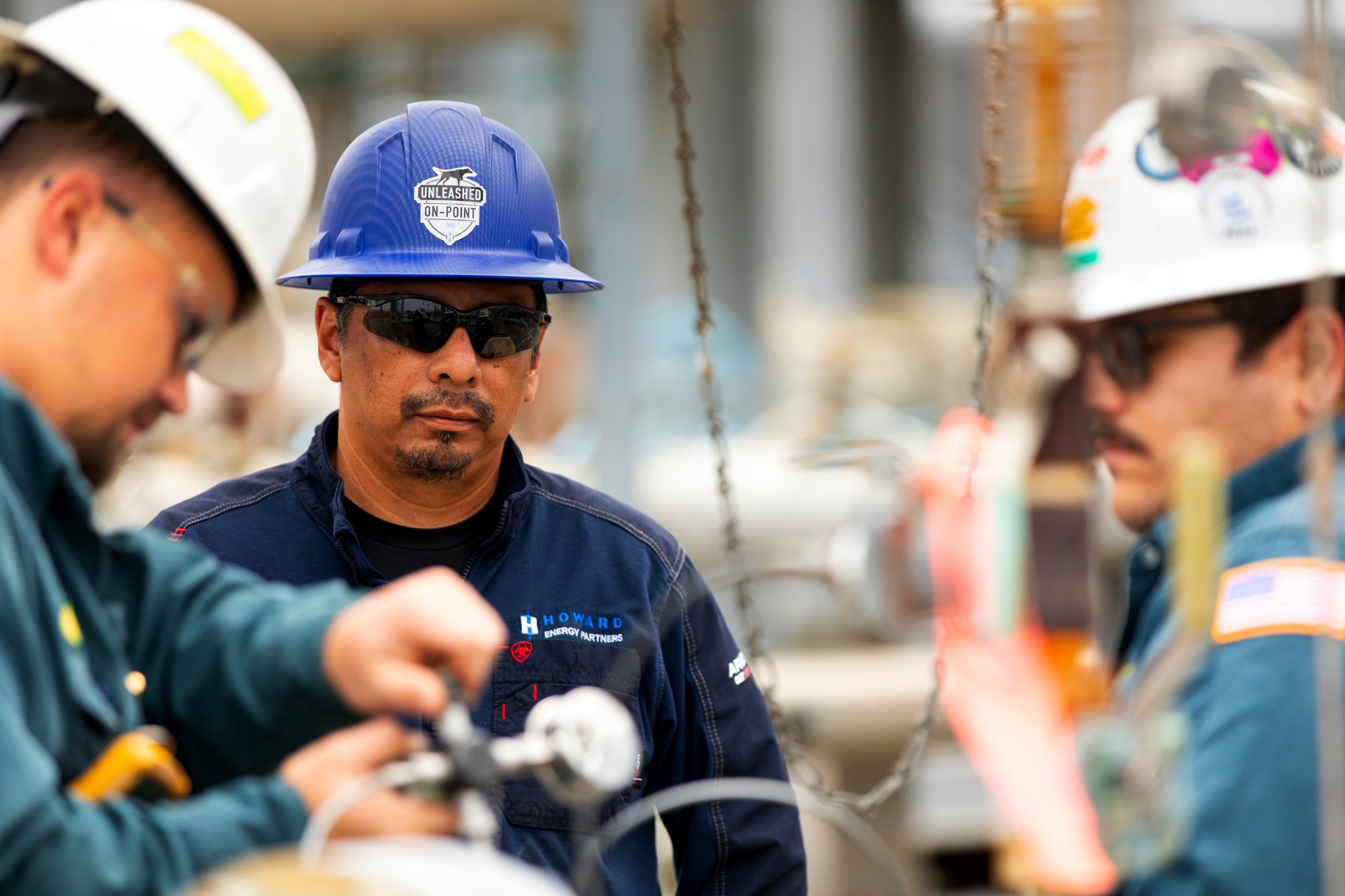 Three men wearing safety helmets and sunglasses working together outdoors with boats in the background.