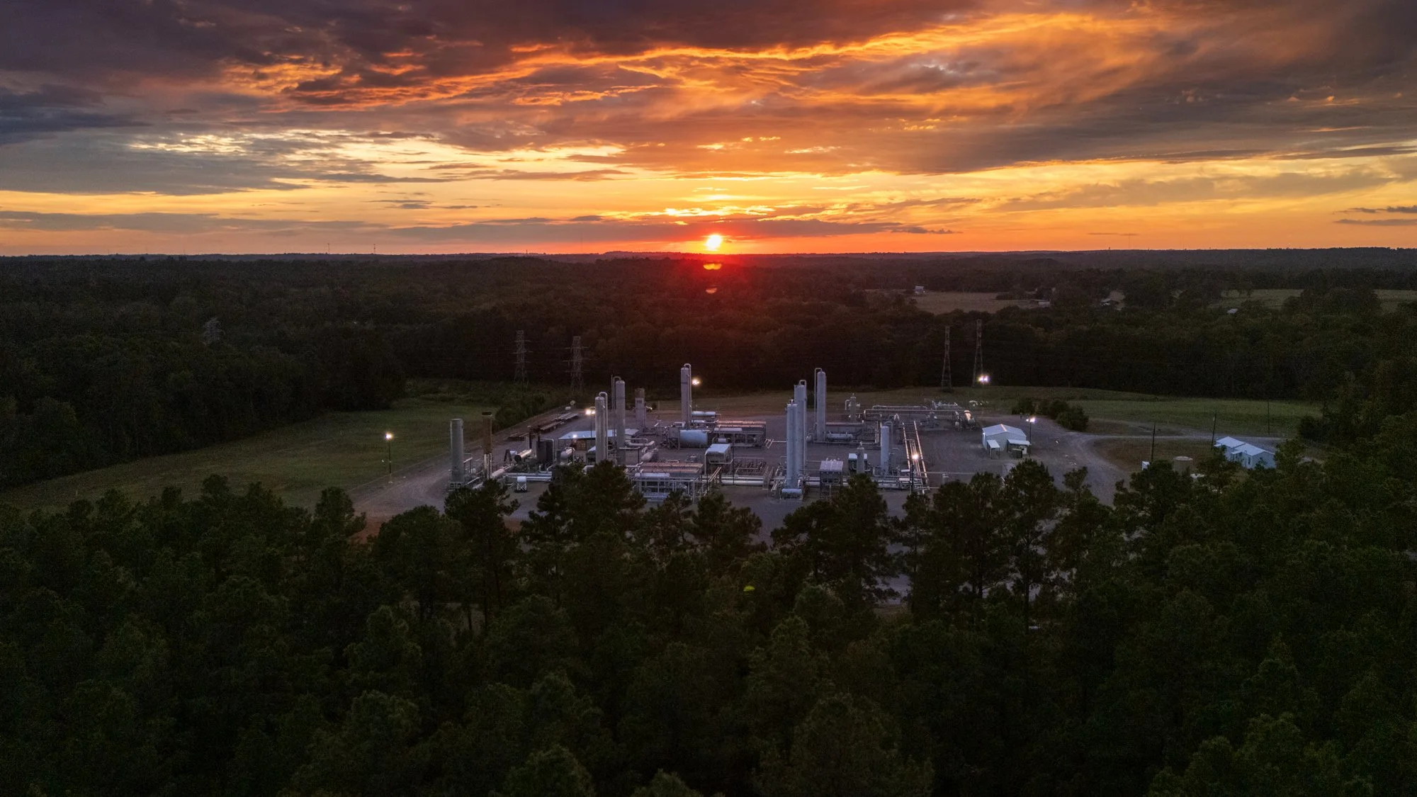 An industrial facility surrounded by a forest, with a sunset sky in the background, featuring clouds and the sun near the horizon.