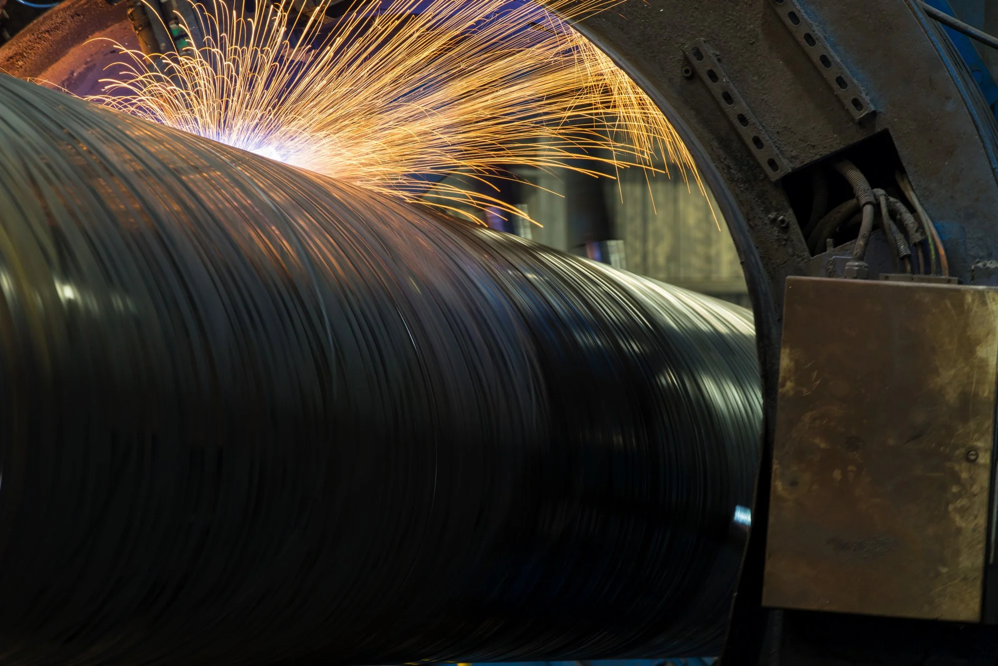 A manufacturing process involving welding or grinding onto a large metal rolled coil, with sparks flying as sparks contact the coil.