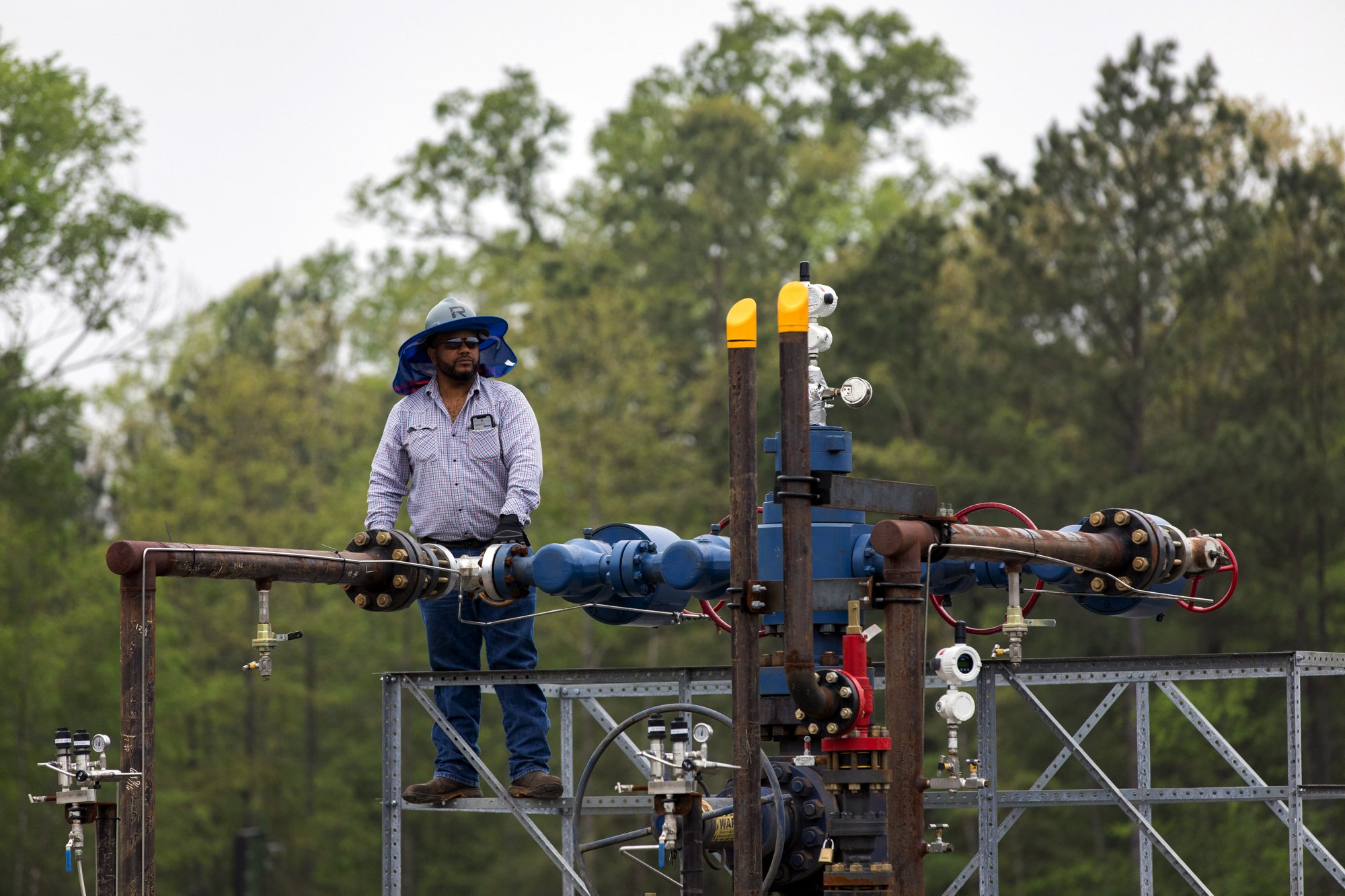Worker standing on a metal platform inspecting large industrial piping with valves and gauges, surrounded by green trees.