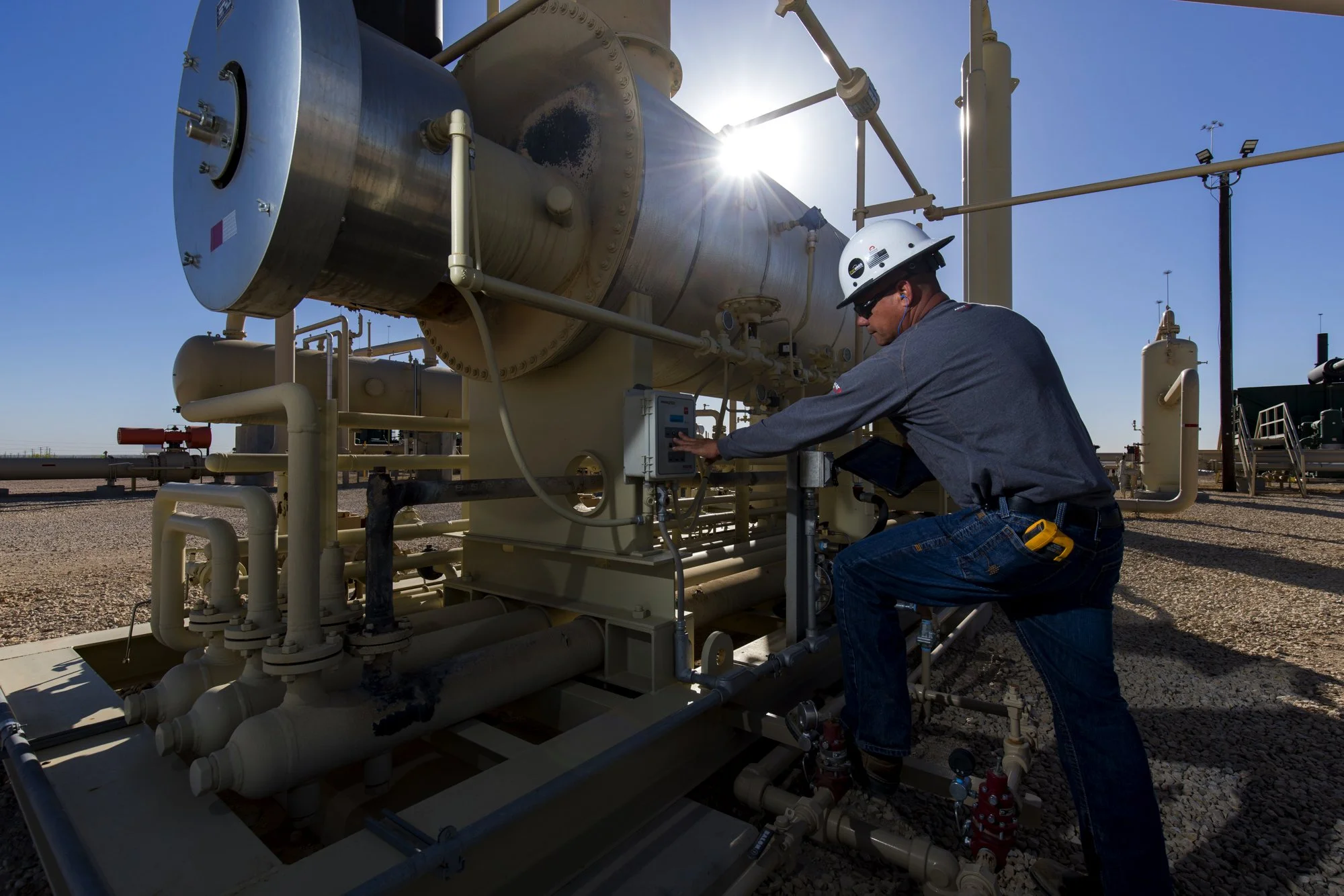 A worker with a white hard hat and sunglasses is operating machinery outdoors, with a bright sun behind the equipment.