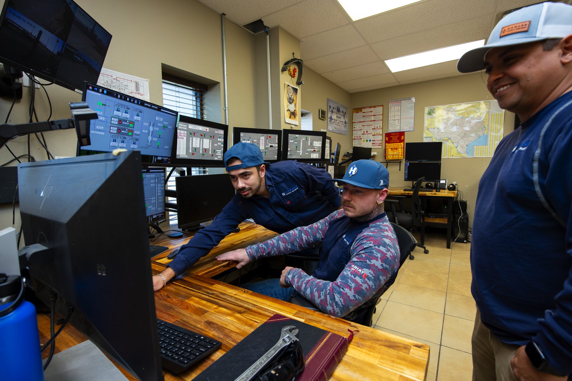 Three people in an operations room looking at multiple computer monitors managing control systems, with one person seated at a desk and the other two standing.