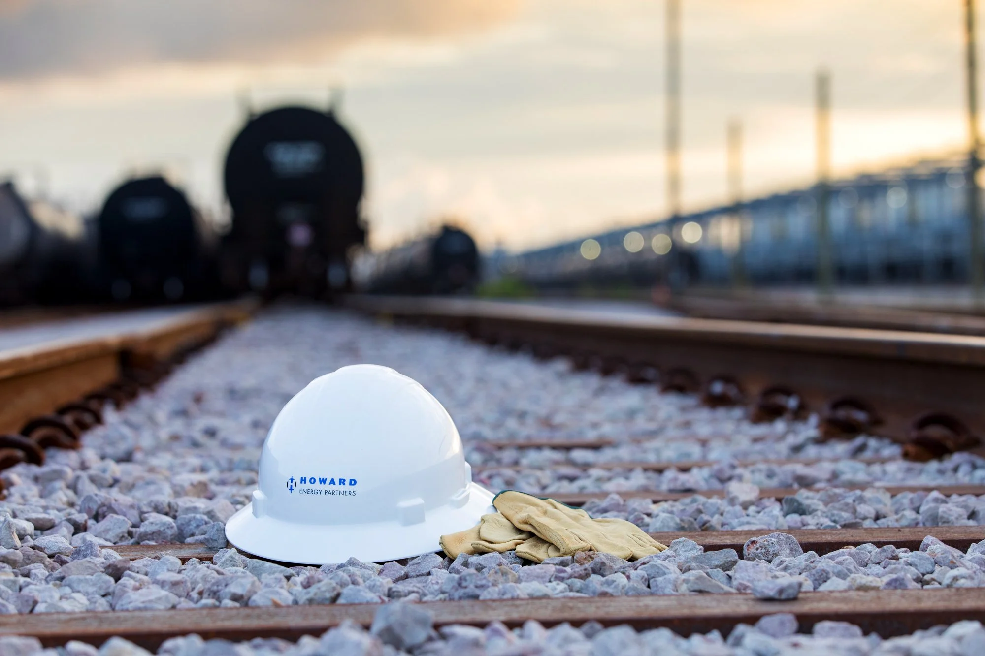 A white hard hat with the logo 'Howard Energy Partners' and yellow work gloves resting on a gravel and wooden railway track, with blurred trains in the background under a cloudy sky.