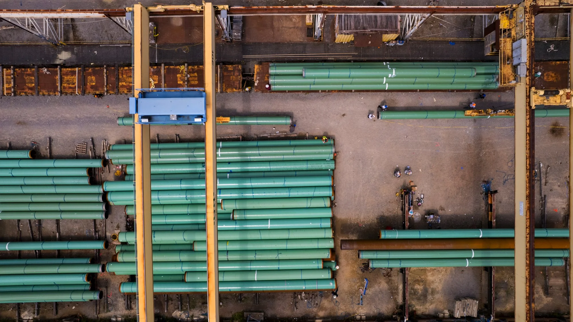 An aerial view of a construction site showing large green pipes, steel structures, construction workers, and various construction equipment.