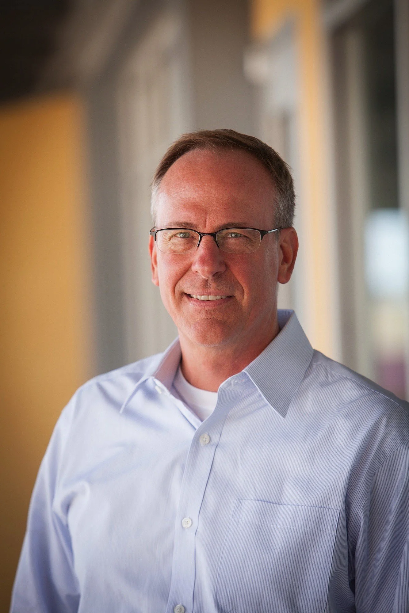A middle-aged man with glasses, short brown hair, wearing a light blue dress shirt, smiling and standing in a warmly lit indoor space with blurred background.