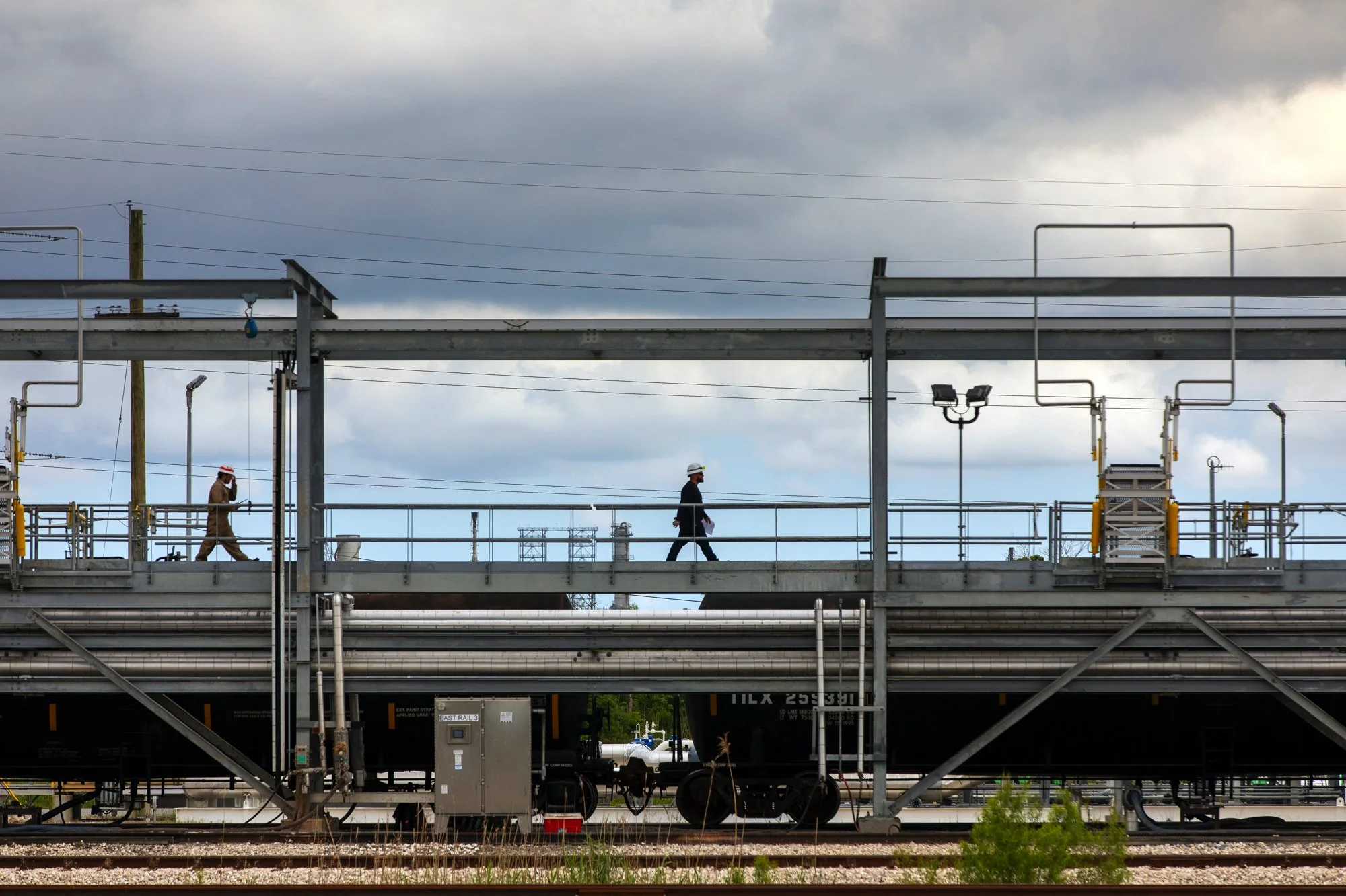 Two people walk along an industrial train bridge under a cloudy sky, with railroad tracks below.