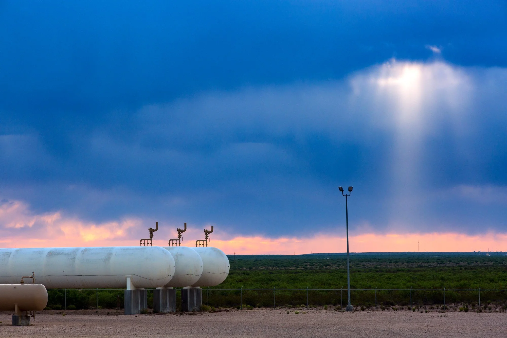 A gas storage tank with pipes and a fence in a desert landscape under a cloudy sky with a ray of sunlight breaking through the clouds.