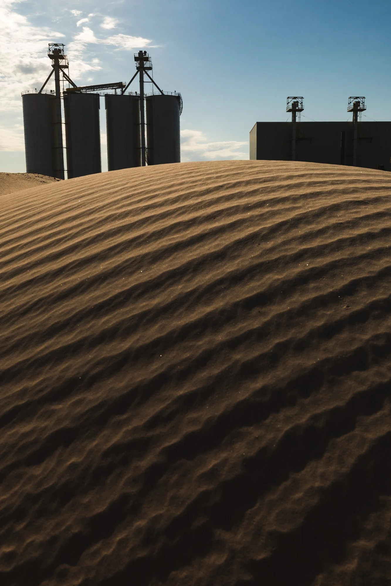 Sand dunes in the foreground with industrial silos and buildings in the background under a partly cloudy sky.