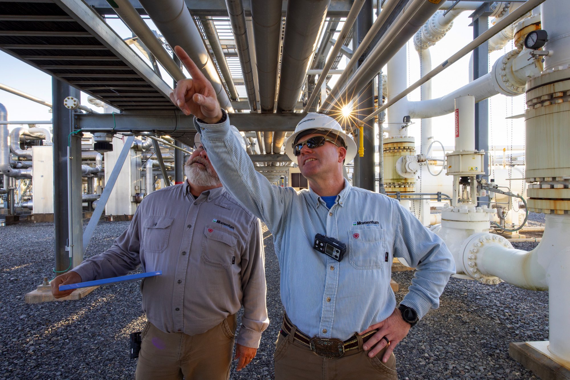 Two male workers in safety gear looking at a device in an industrial facility with pipes and machinery.