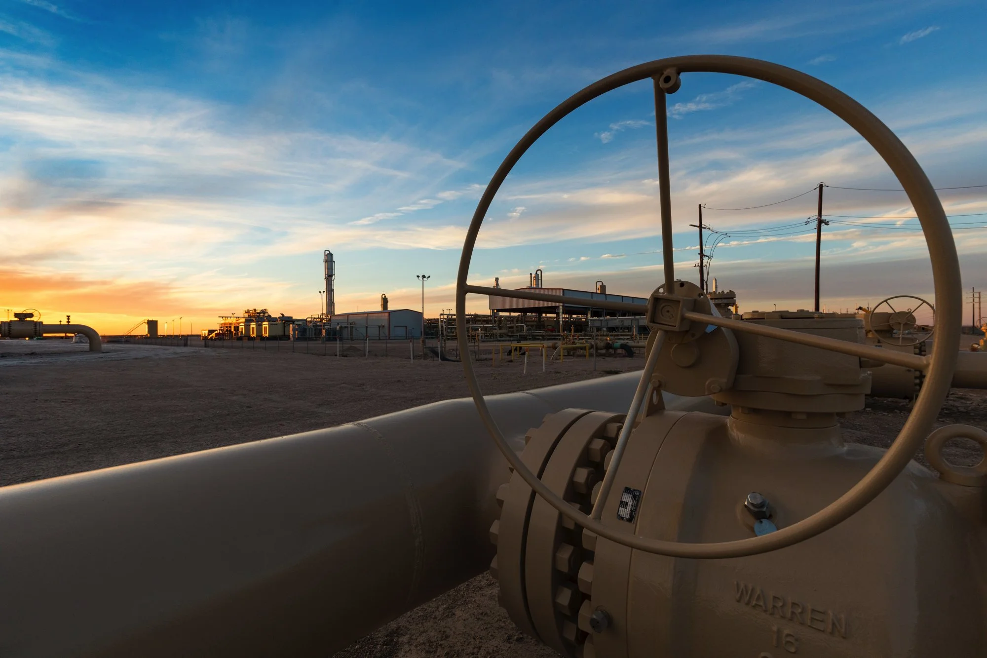 Close-up of industrial pipe fitting at sunset in an oil or chemical refinery, with a background of equipment, pipelines, and utility poles under a partly cloudy sky.