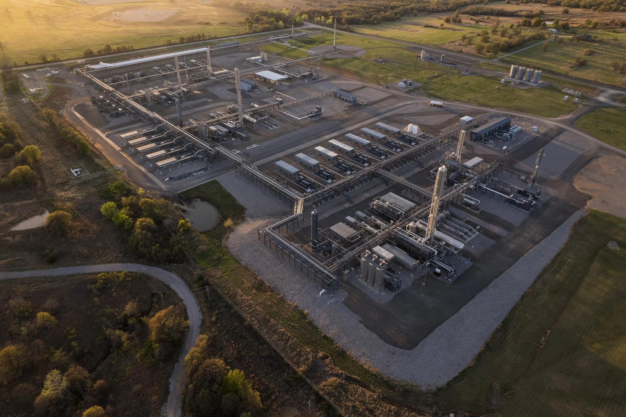 Aerial view of an industrial oil or gas processing facility with pipelines, storage tanks, and equipment, surrounded by fields and greenery at sunset.
