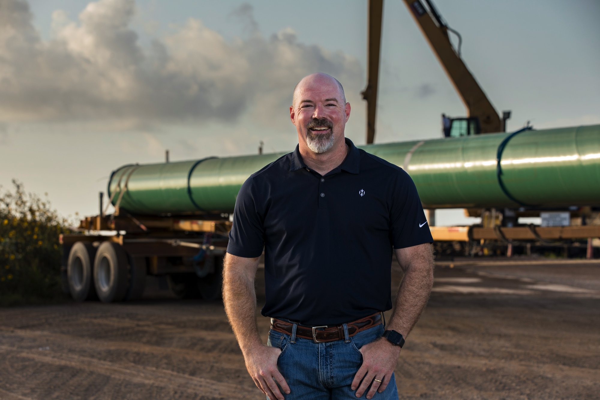 A man standing outdoors in front of a large green industrial pipe on a trailer, with construction equipment in the background. The man is smiling, wearing a black polo shirt, jeans, and a watch.