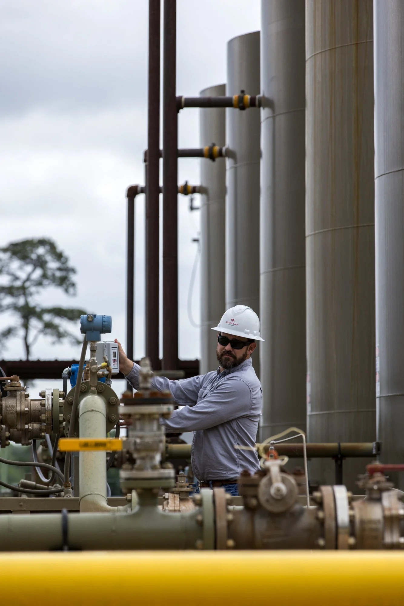 A man wearing a hard hat and sunglasses working with industrial equipment outdoors, with large tanks in the background.