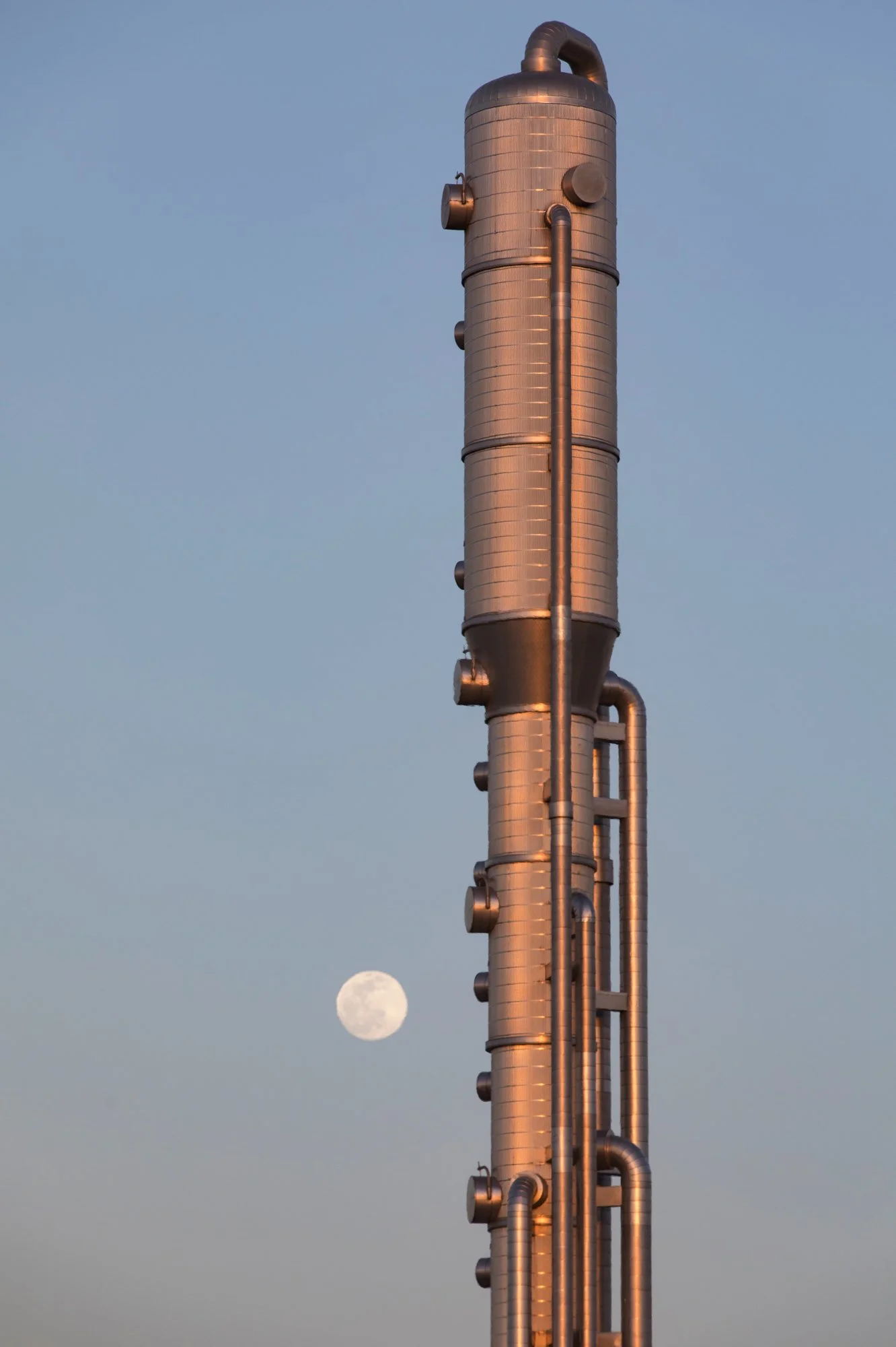 A tall industrial chimney with pipes attached, set against a clear sky during sunset, with the moon visible in the background.